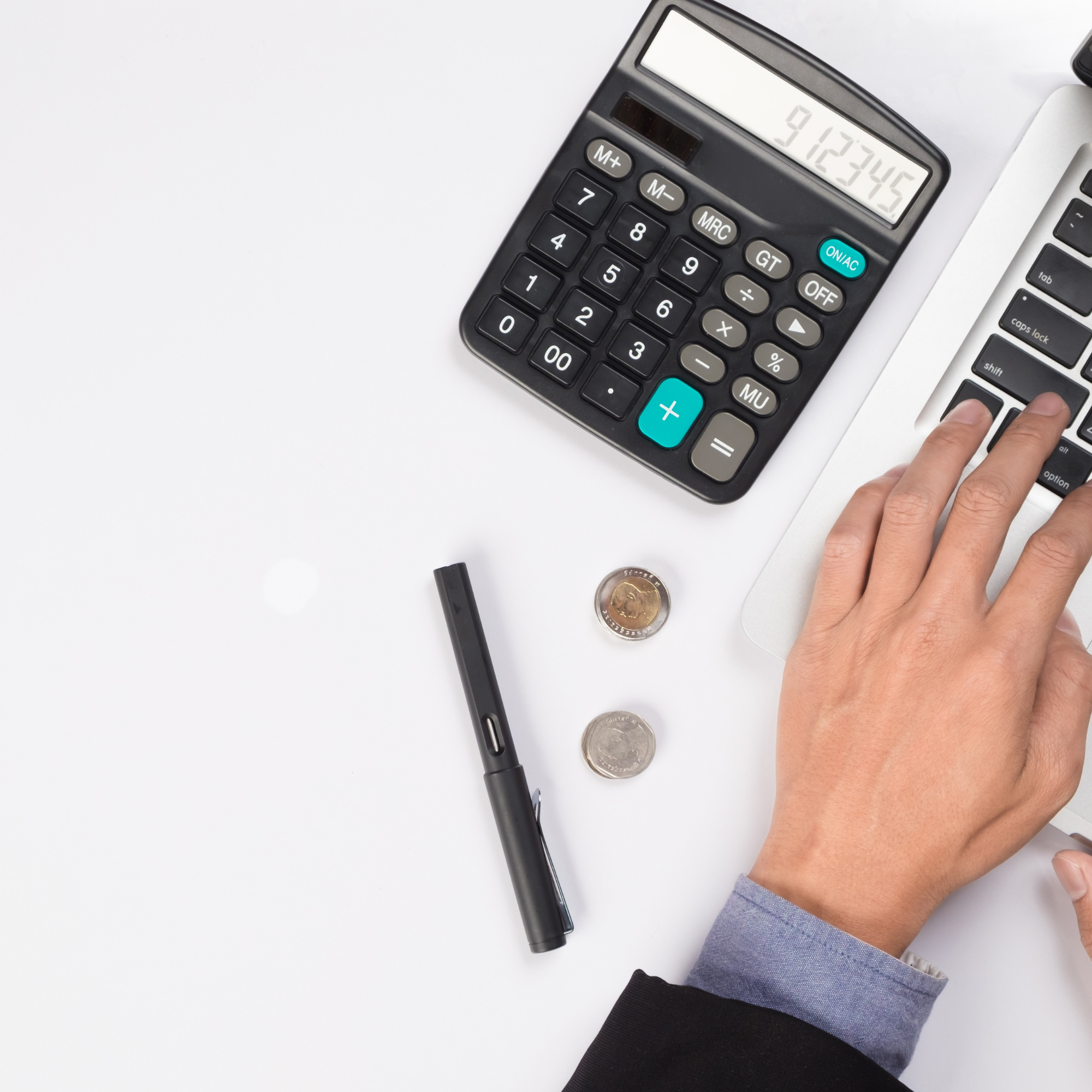 Close-up of a calculator showing numerical results, a person's hand typing on a laptop keyboard, a pen, and coins on a white desk.