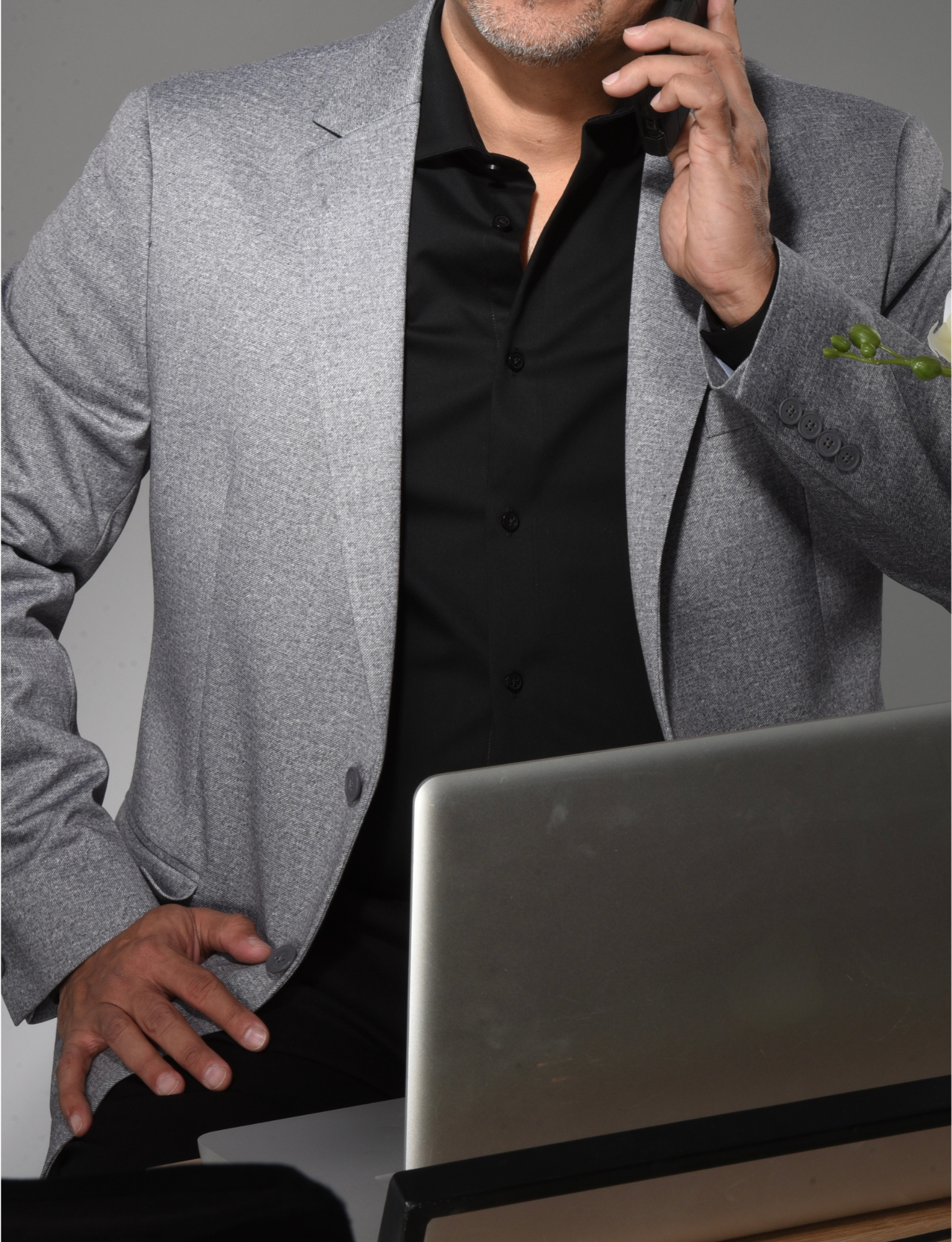 A man wearing a gray blazer and black shirt, talking on a cell phone while seated at a desk with a laptop computer.
