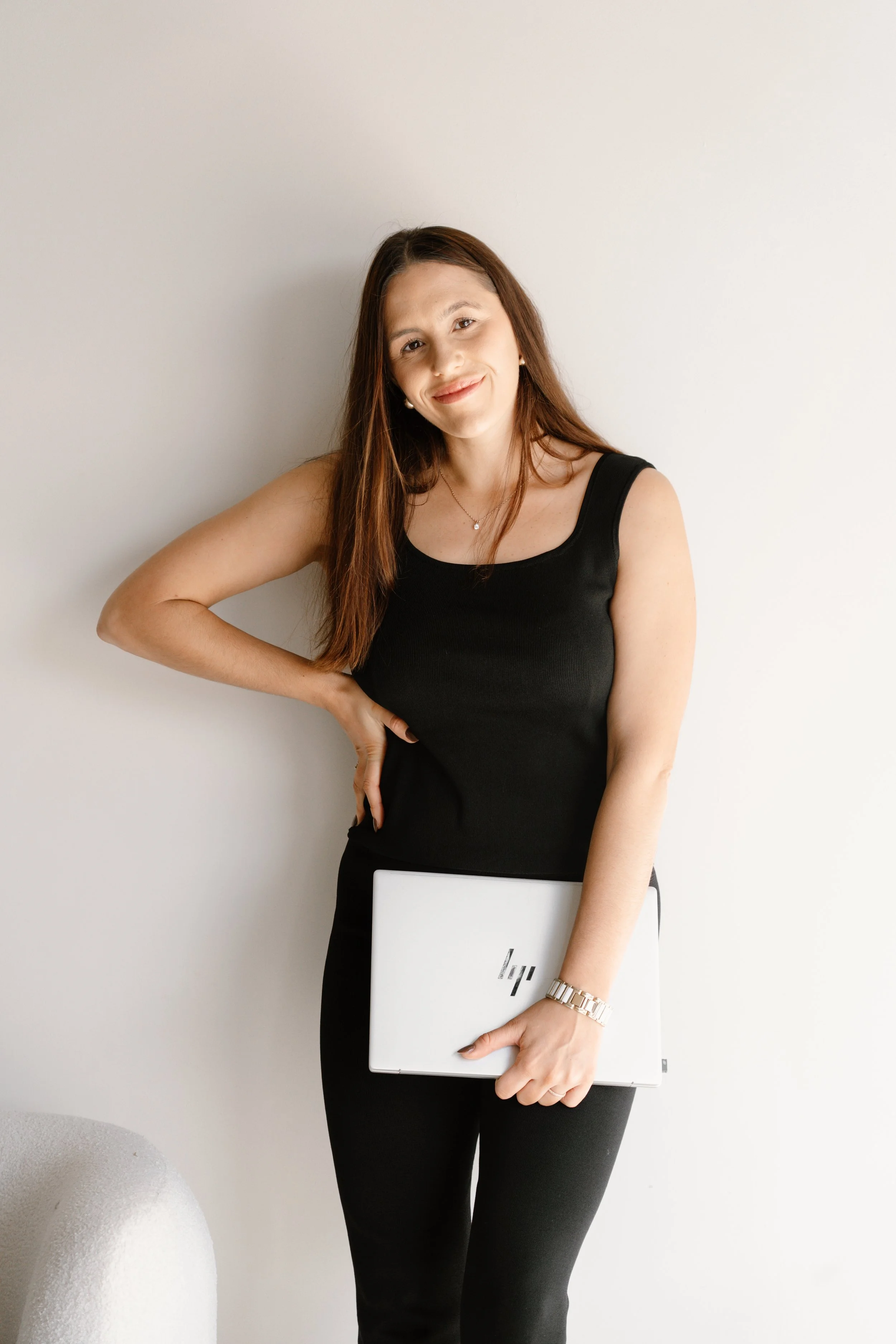 A woman with long brown hair wearing a black sleeveless top and black pants, holding a silver HP laptop, standing against a plain white wall.