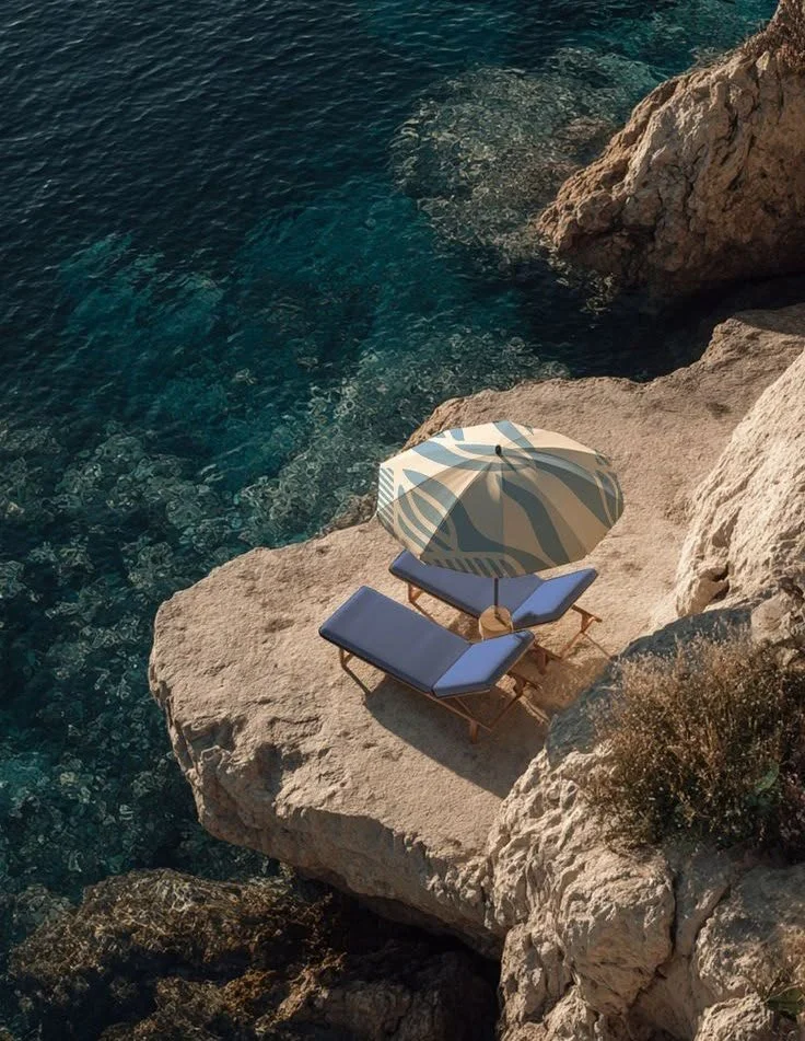 A rocky coastline with two blue lounge chairs, a striped beach umbrella, and a small table on a large flat rock near the water.