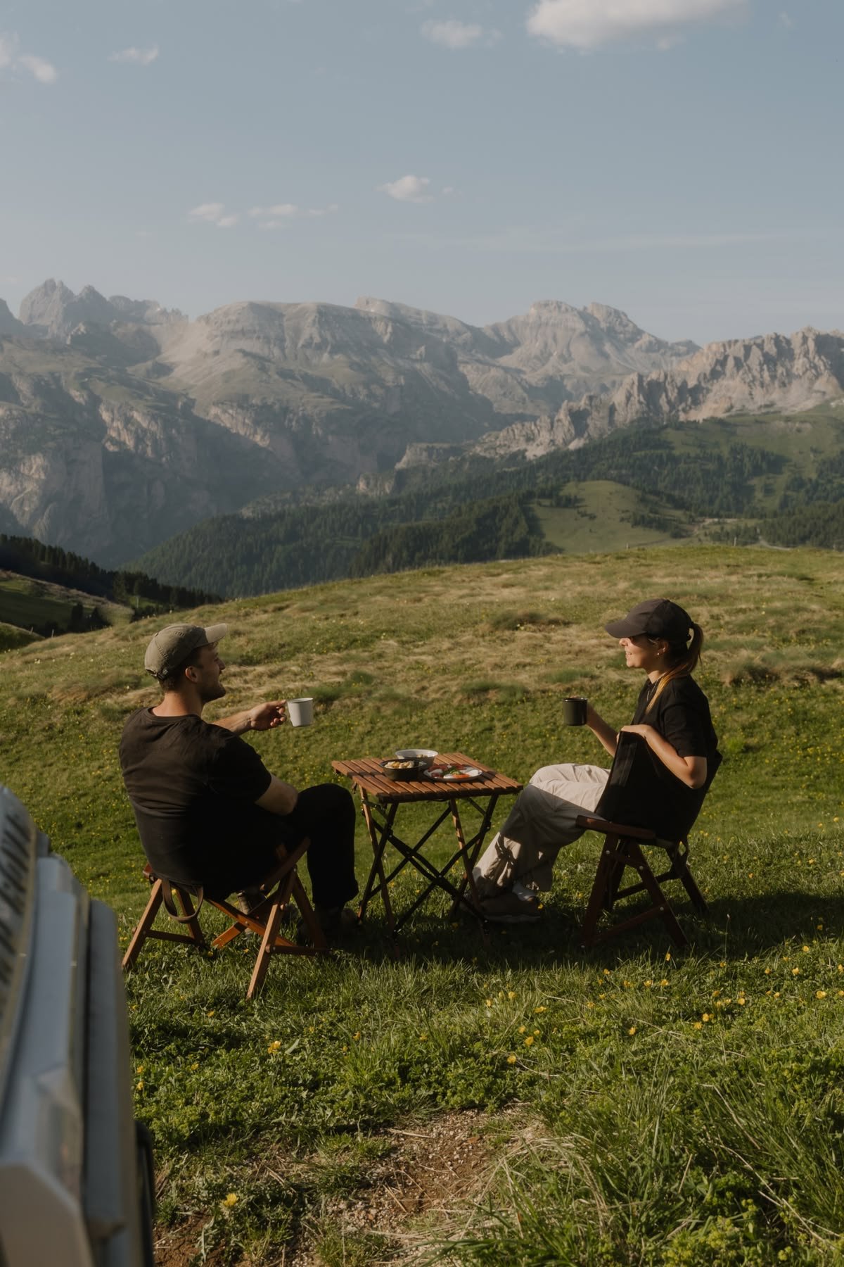 Two people sit on foldable chairs, having coffee at a small wooden table in a grassy field with mountains in the background, under a partly cloudy sky.