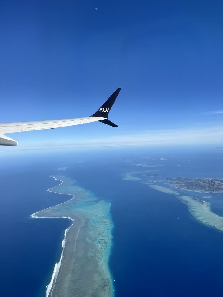 Aerial view from an airplane window showing a reef and islands below the clear blue ocean, with the wing marked 'FIJI' visible.