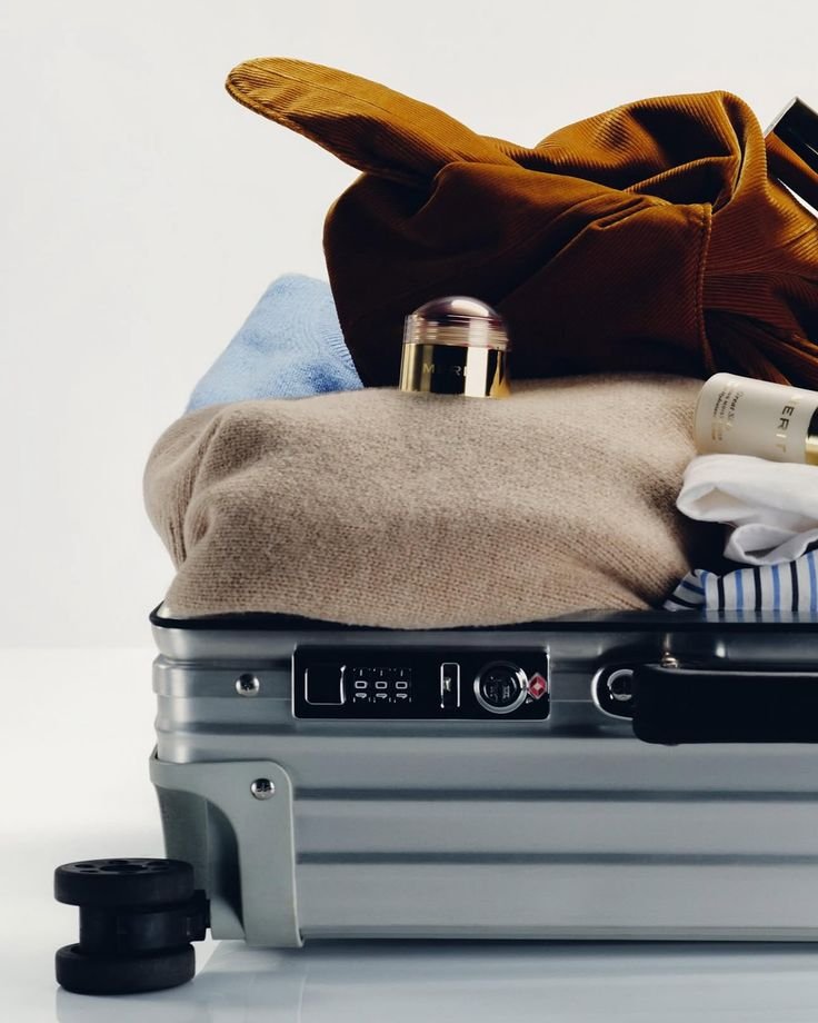 Open silver suitcase filled with clothes and personal items, with some items spilling out, placed on white surface.