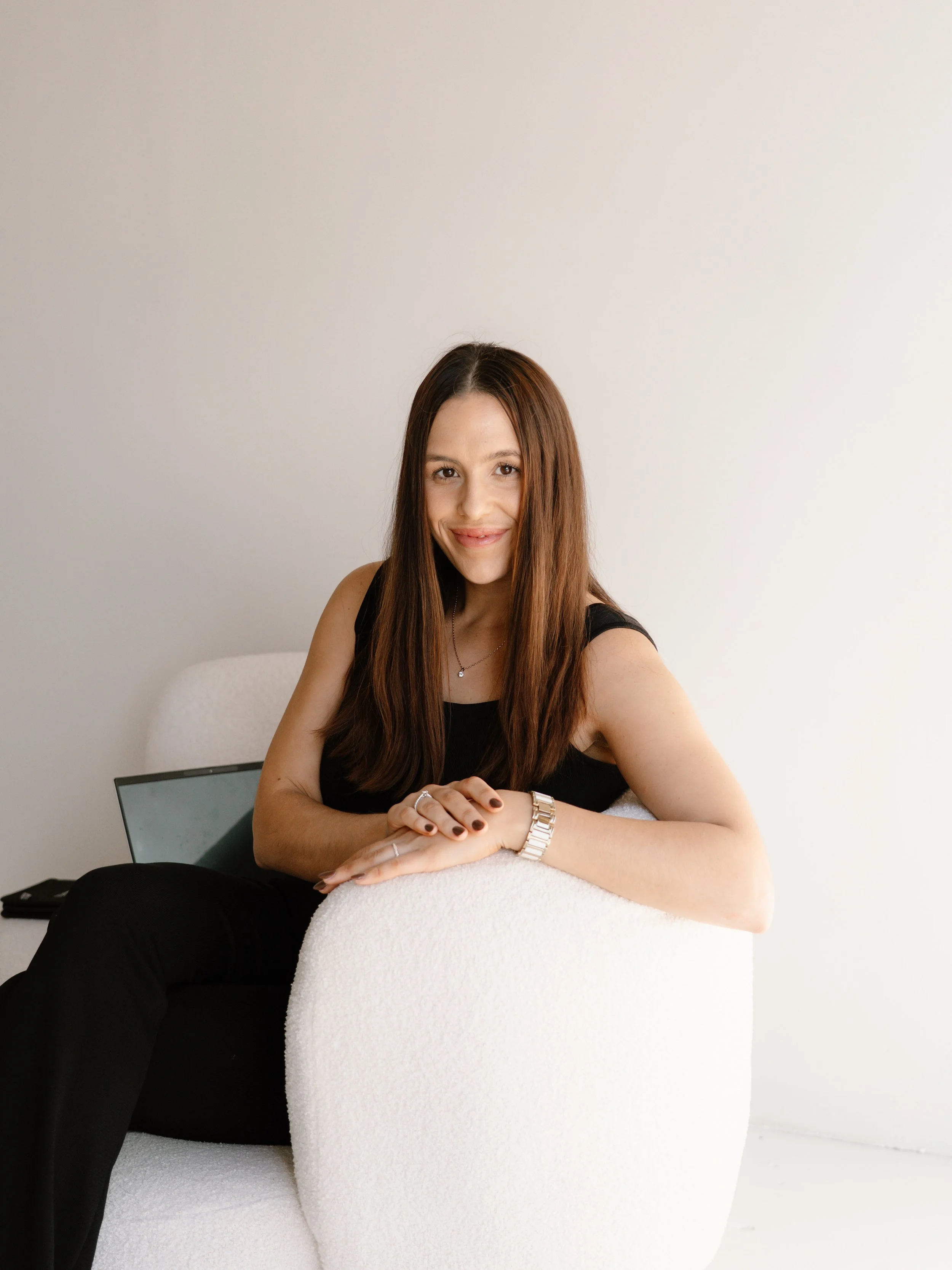 A woman with long brown hair, wearing a black sleeveless top, sitting on a white textured armchair, smiling and looking at the camera.