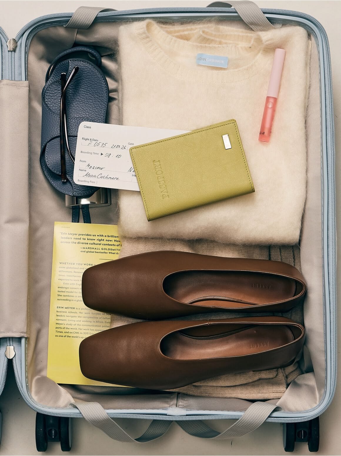 Open suitcase containing a pair of brown shoes, a yellow passport holder, a cream sweater, a pink lip gloss, a black eyeglasses case, a flight ticket, and a yellow informational paper.