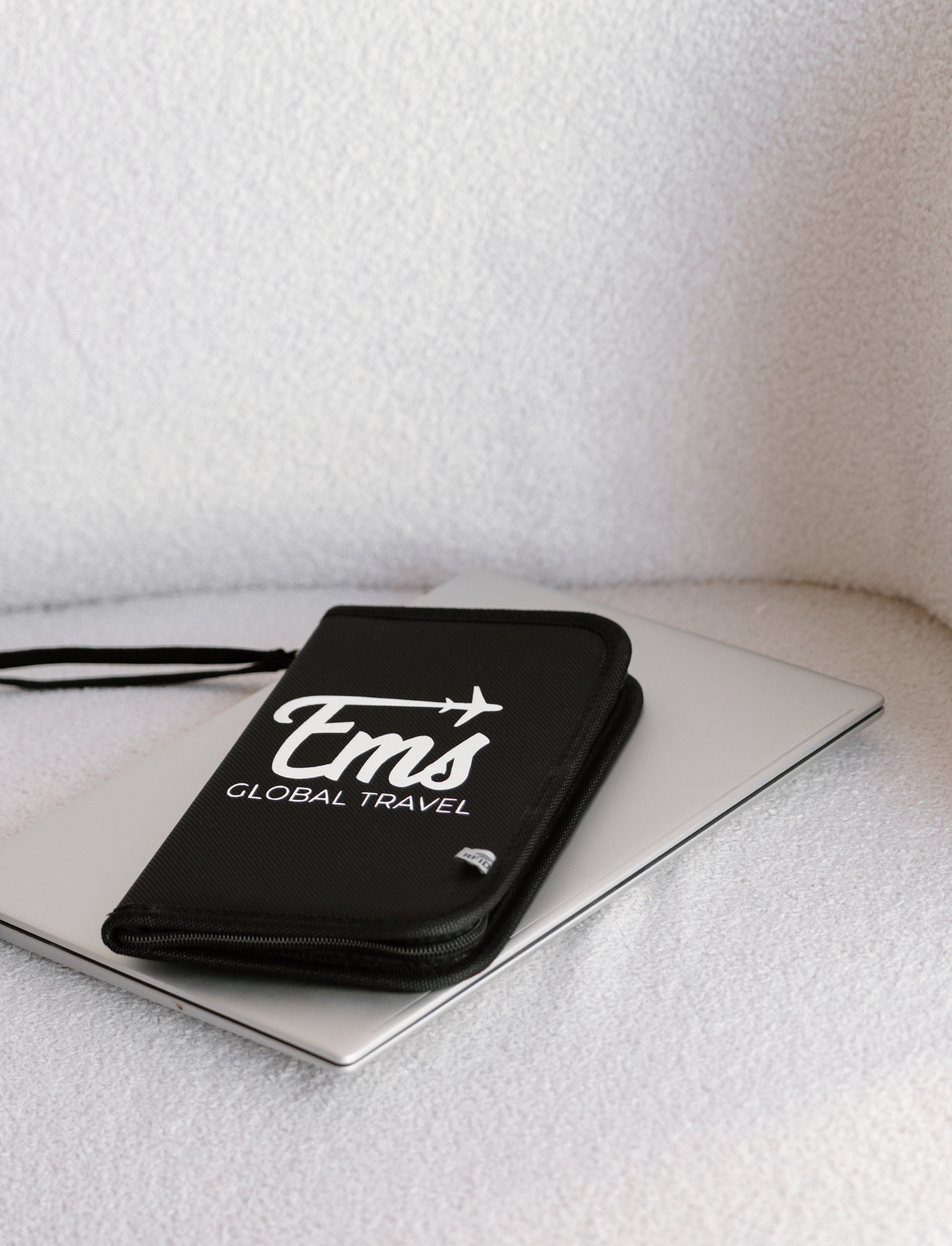 A black travel wallet with the Tim's Global Travel logo placed on a closed silver laptop on a textured white surface.