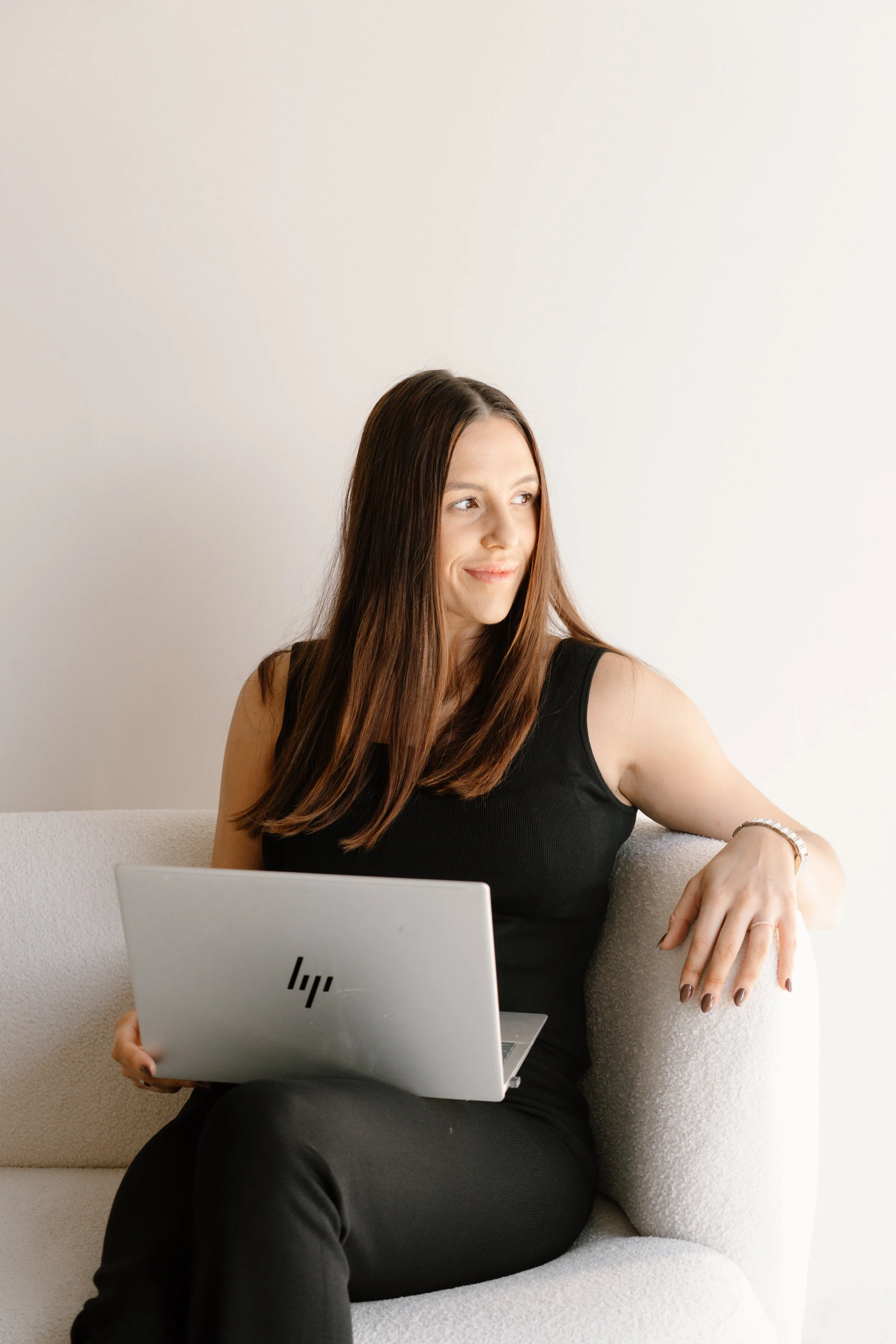 A woman sitting on a beige couch with a laptop, looking to her left with a slight smile.