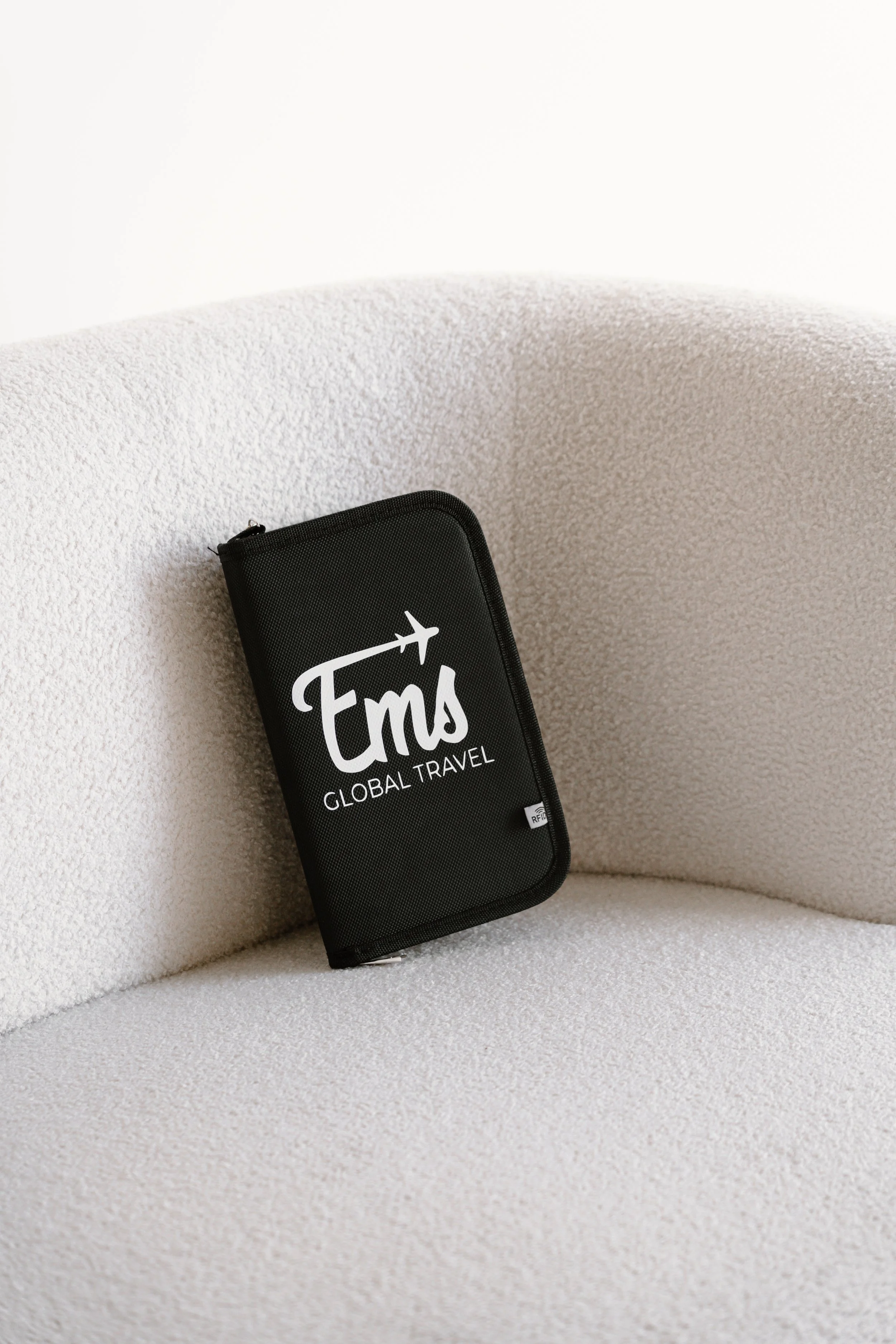 Close-up of a black travel wallet with white text that says "Em's Global Travel" and an airplane graphic, resting on a textured white curved surface.