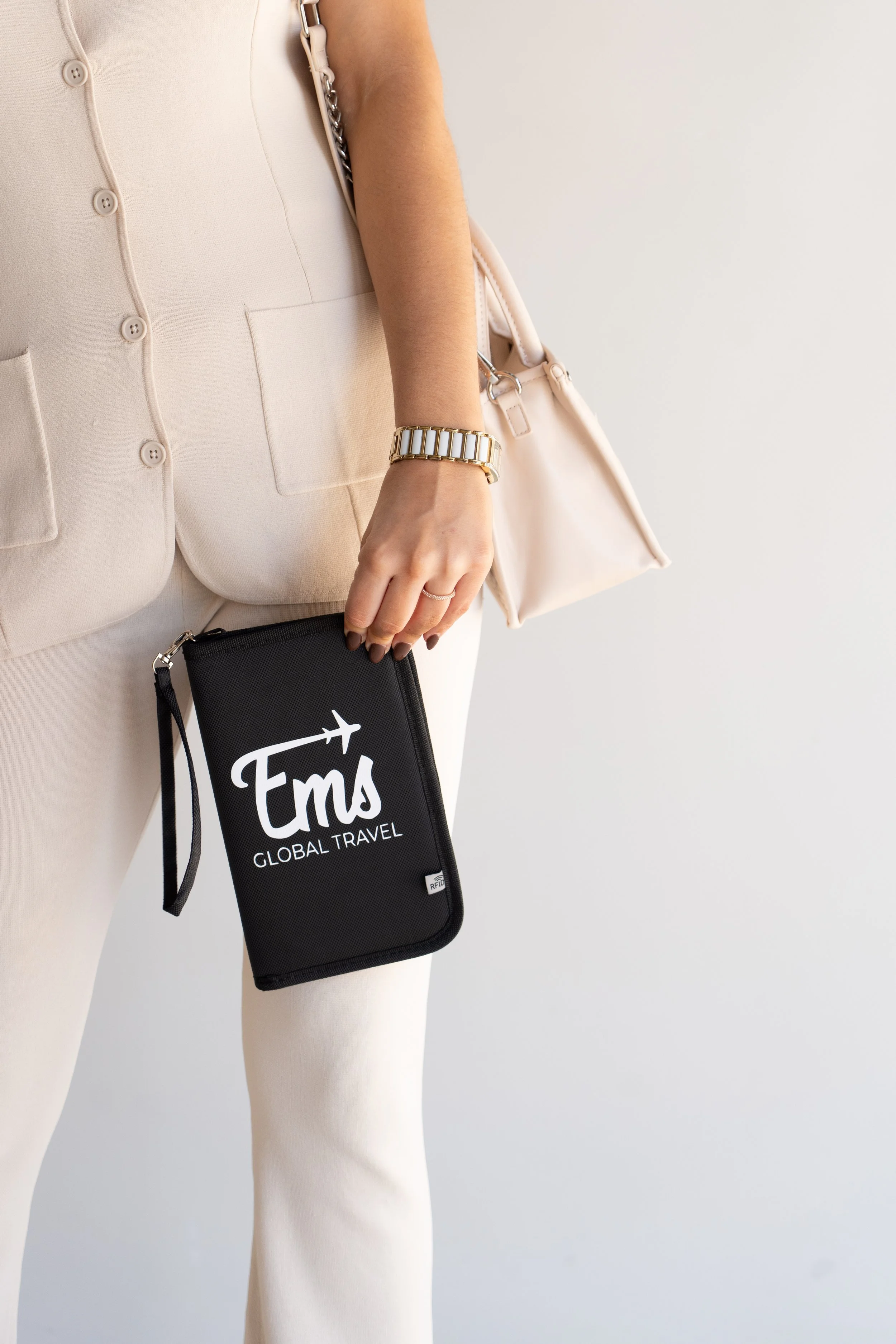 Close-up of a woman's torso wearing a beige blazer, holding a black travel pouch with 'FMS' and 'Global Travel' printed on it, and carrying a beige handbag.