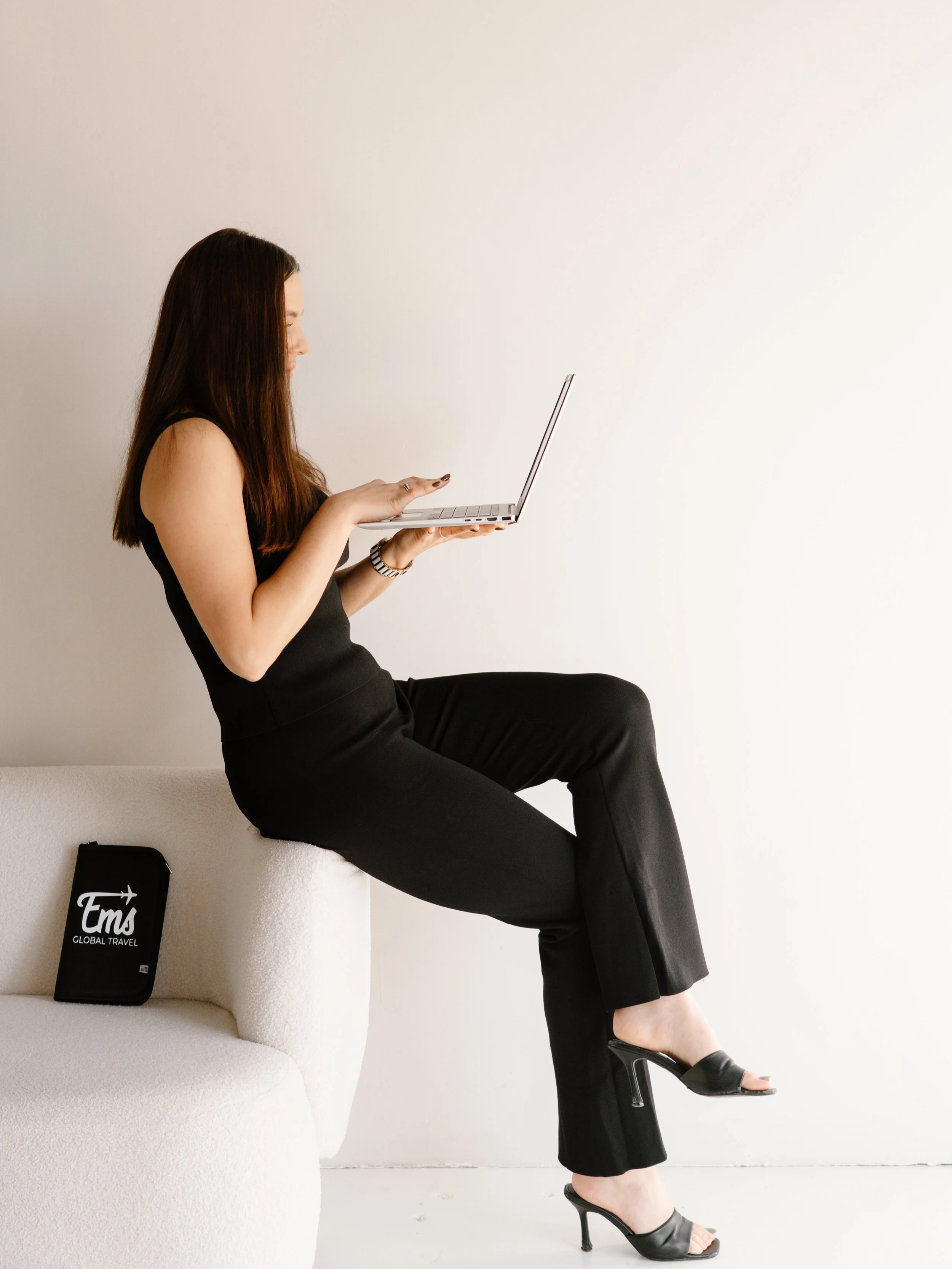 A woman wearing black pants and heels is sitting on a white sofa, looking at a laptop held in her hands. There is a black travel pouch with white text reading 'Em's GLOBAL TRAVEL' on the sofa.