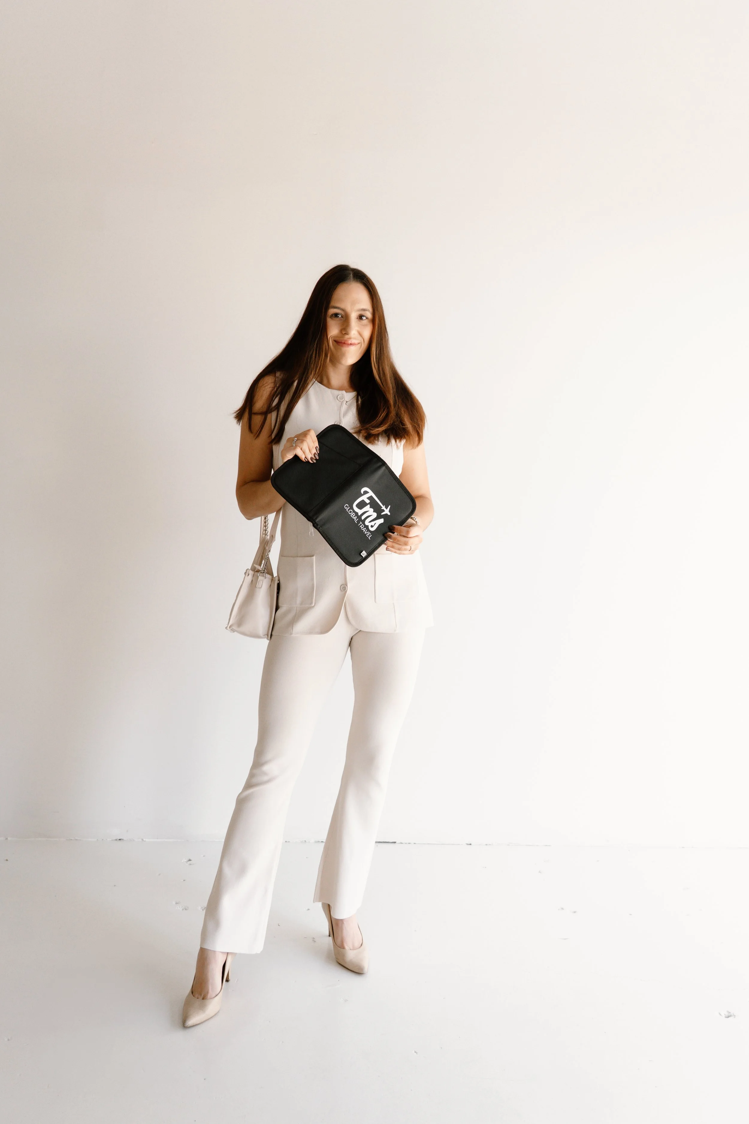 A woman with long brown hair wearing a white sleeveless top, white pants, and beige high heels, standing against a plain white wall. She is holding a black travel pouch with the logo 'Fly' and the text 'Global Travel.' She is also carrying a white shoulder bag.