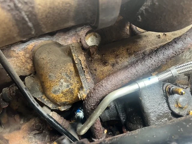 Close-up view of a rusty, aged engine part with a metal tube attached, surrounded by corroded metal surfaces and components.