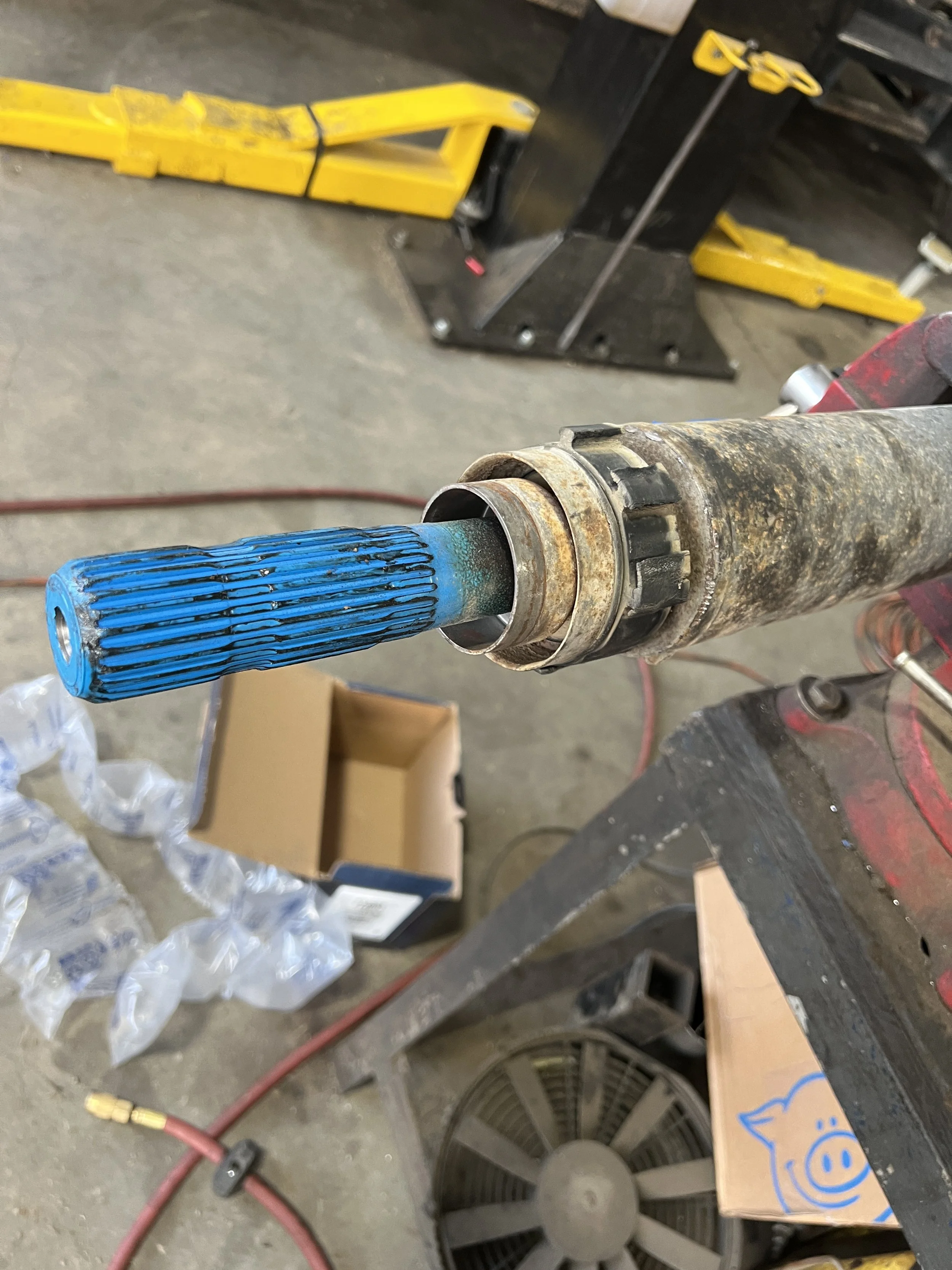 Close-up of a rusty driveshaft with a blue protective cover at the end, placed on a workbench in a garage or workshop with tools and equipment in the background.