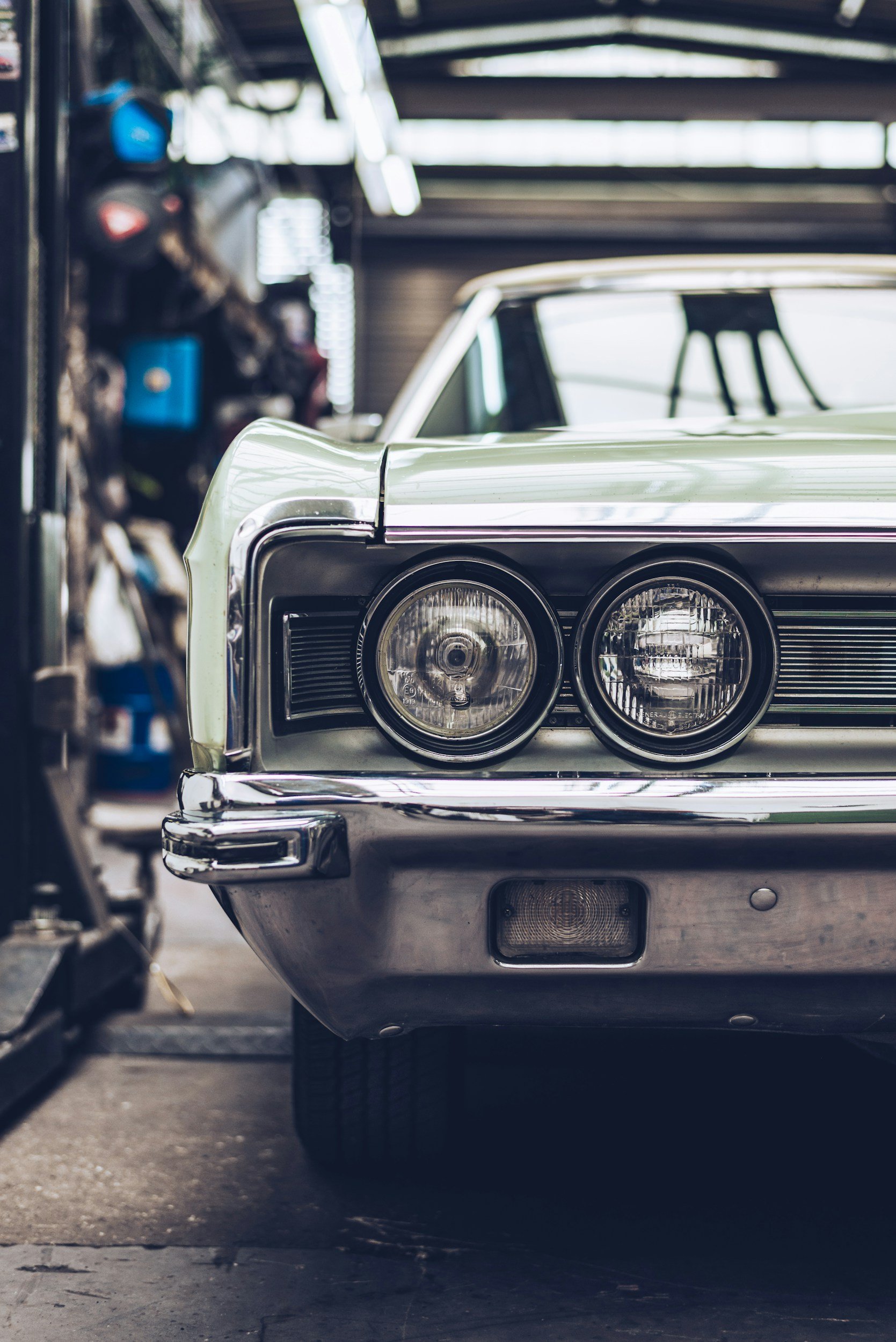 Close-up of the front end of a vintage car, showing its headlights, grille, and bumper, in a garage or workshop setting with tools and equipment visible in the background.