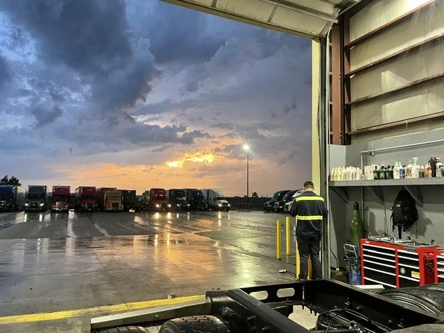 View of a parking lot seen from inside a service garage, with trucks parked in the distance and a sunset sky with dark clouds. A man in a reflective jacket faces away, standing near shelves and tools.
