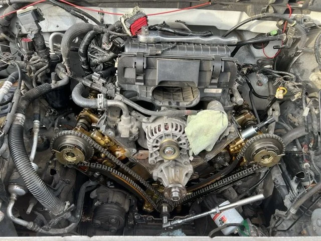 Close-up view of a car engine with timing belts, pulleys, and various engine components visible. The engine appears to be in the process of repair or maintenance.