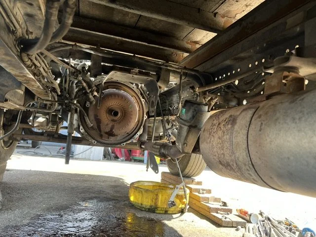 Underneath a vehicle, showing engine components and a large exhaust pipe, with an oil drain pan placed on the ground.