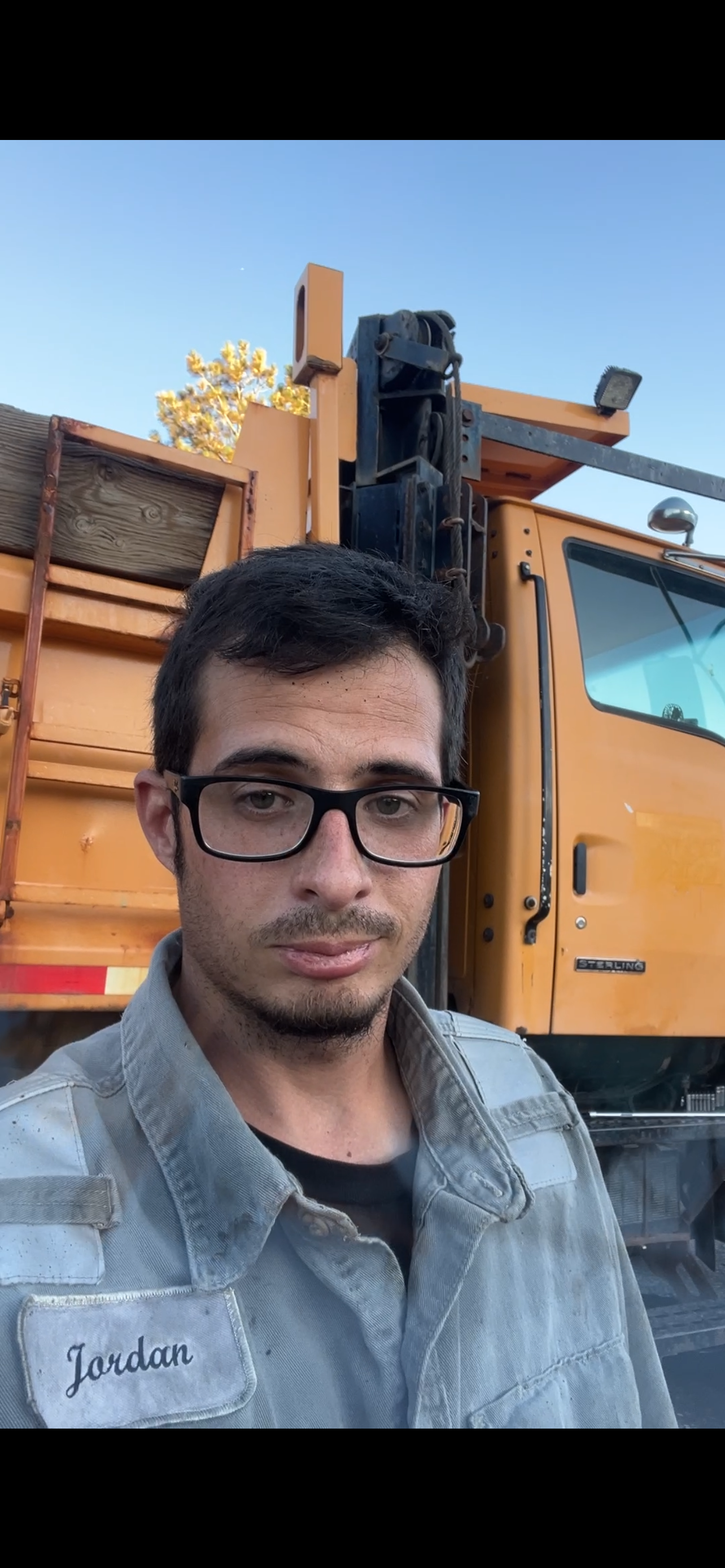 Self-portrait of a man with glasses and a gray shirt in front of orange construction or utility truck with wooden panels and equipment.
