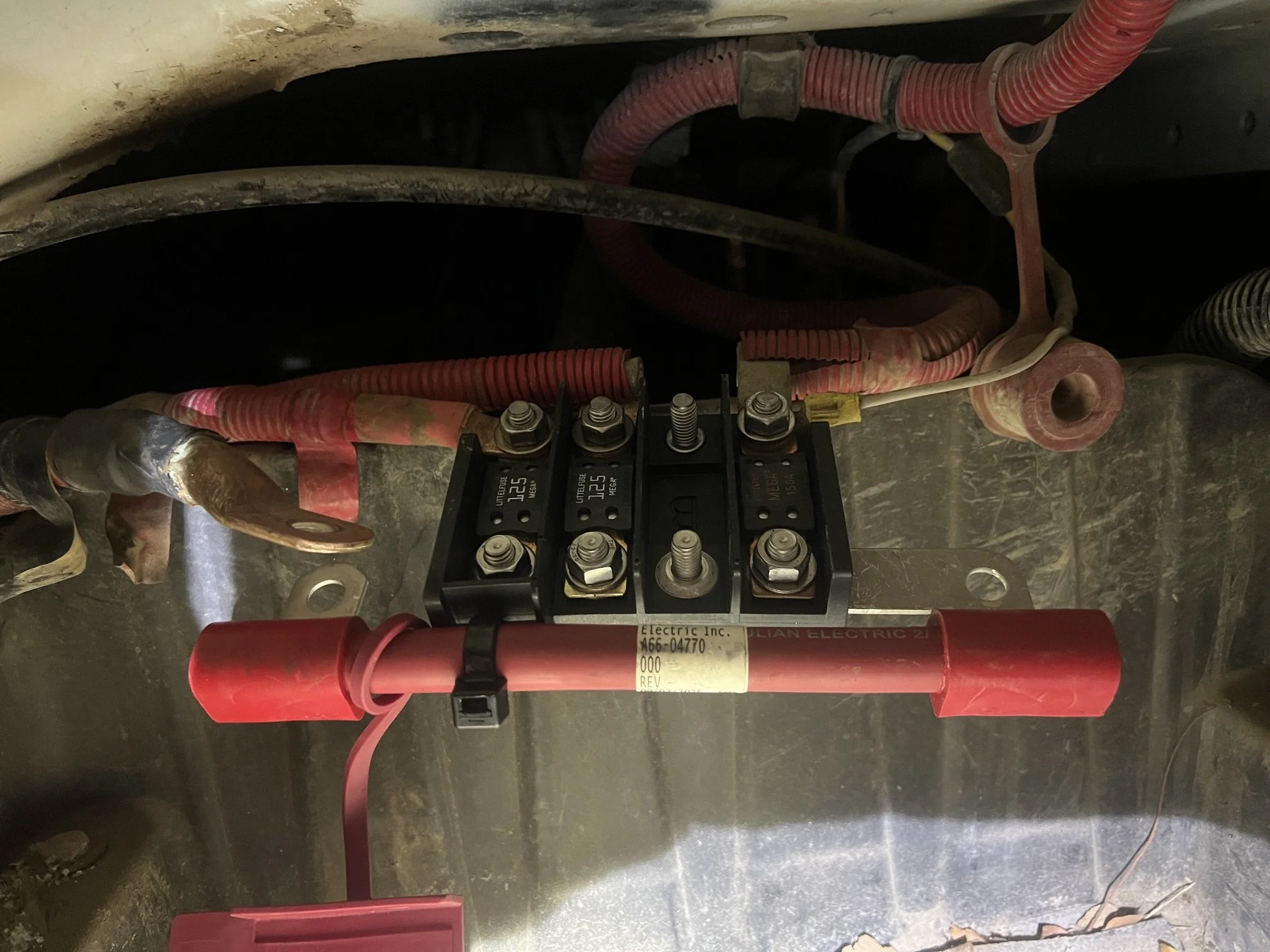 An electrical system with red and gray cables connected to a black terminal block, mounted on a metallic surface, with various bolts and a white sticker with part numbers.