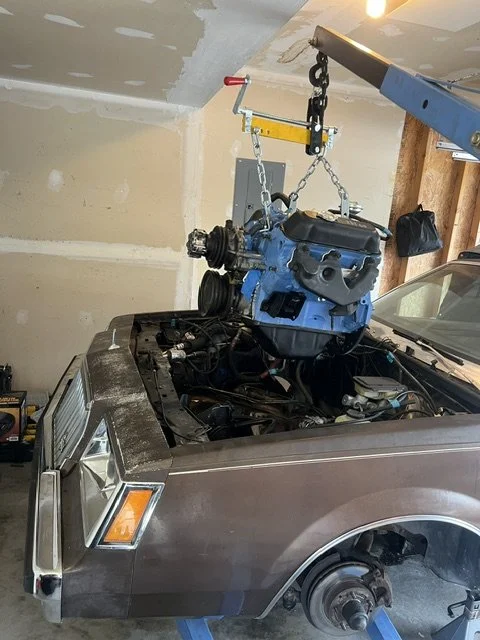 A blue engine is being lifted out of a vintage brown car's engine bay using a hoist in a garage.