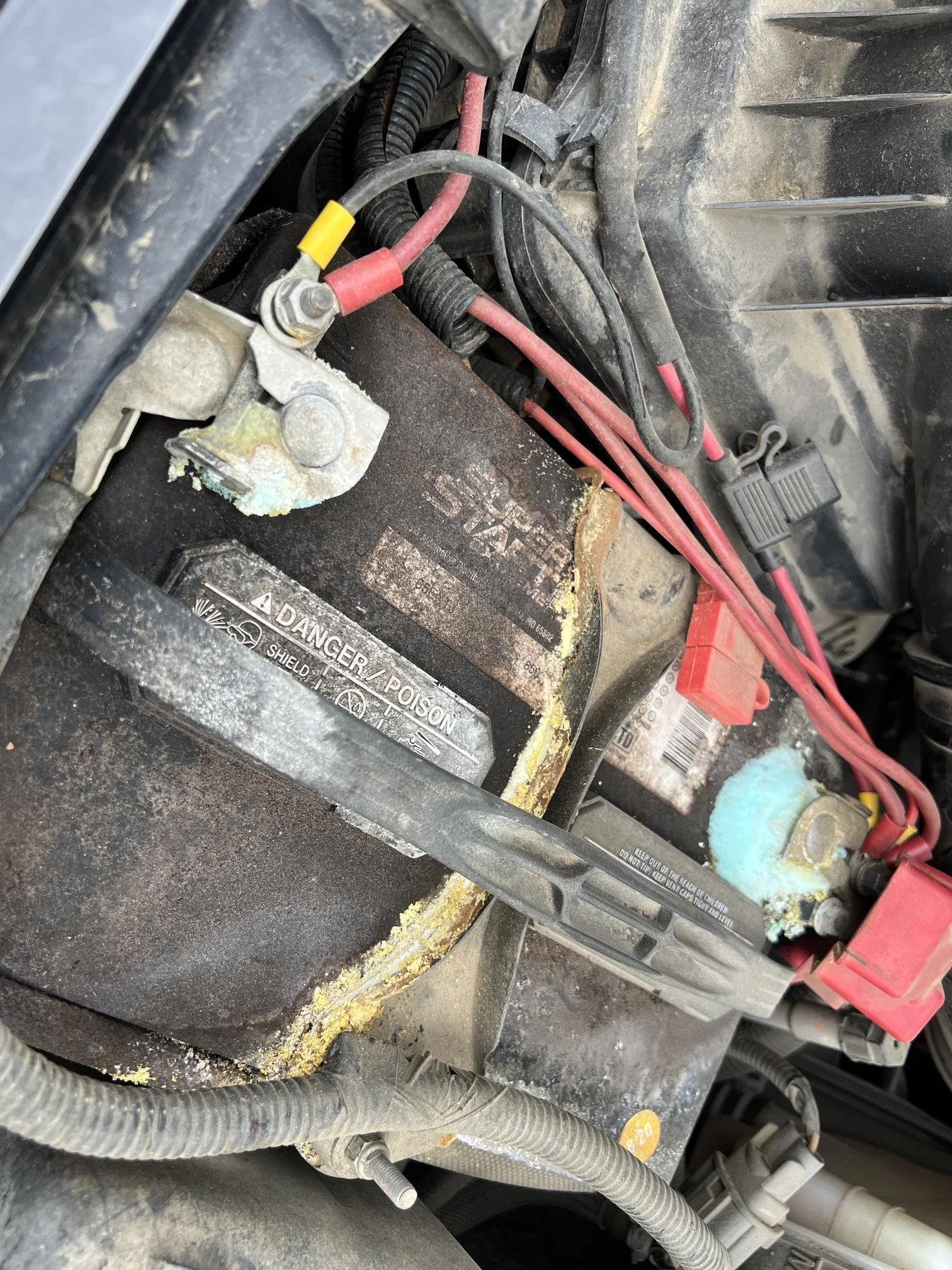 Close-up view of a car battery with red and black cables connected, located inside a dirty engine compartment with wires and hoses nearby.