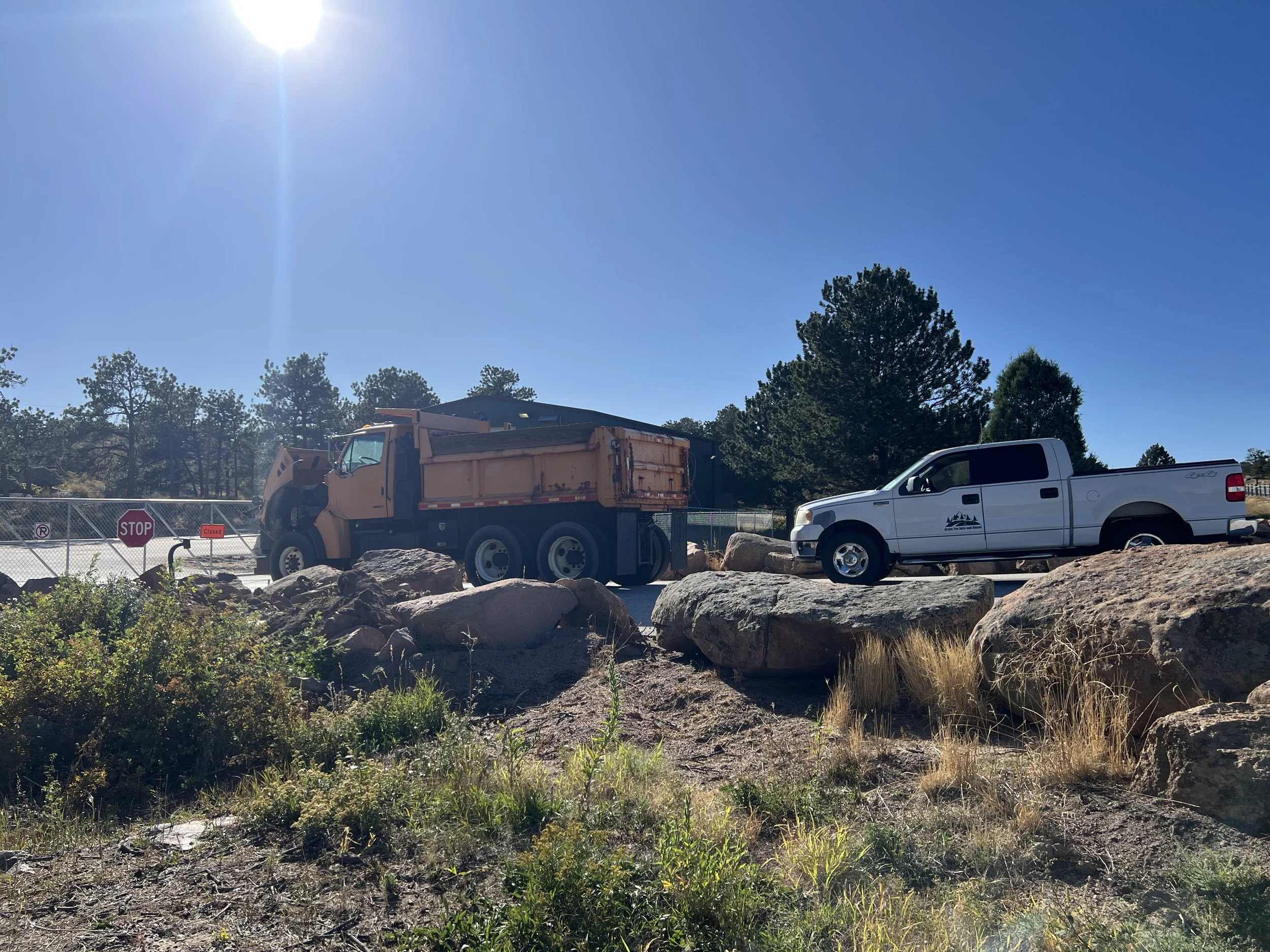 A street view with construction vehicles, including a yellow dump truck and a white pickup truck, surrounded by rocks, rocks, trees, a fence, and clear blue sky with the sun shining.