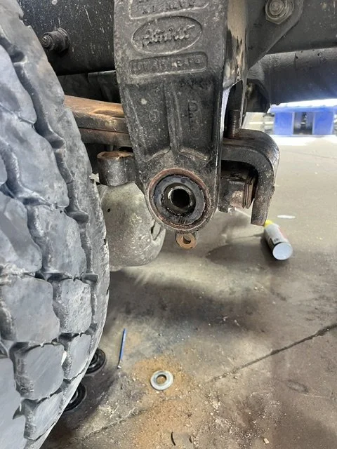 Close-up of a vehicle's rear hitch or towing assembly with a missing or removed part, labeled 'Pintle Hook', showing rust and dirt, on a concrete garage floor with tools and debris nearby.