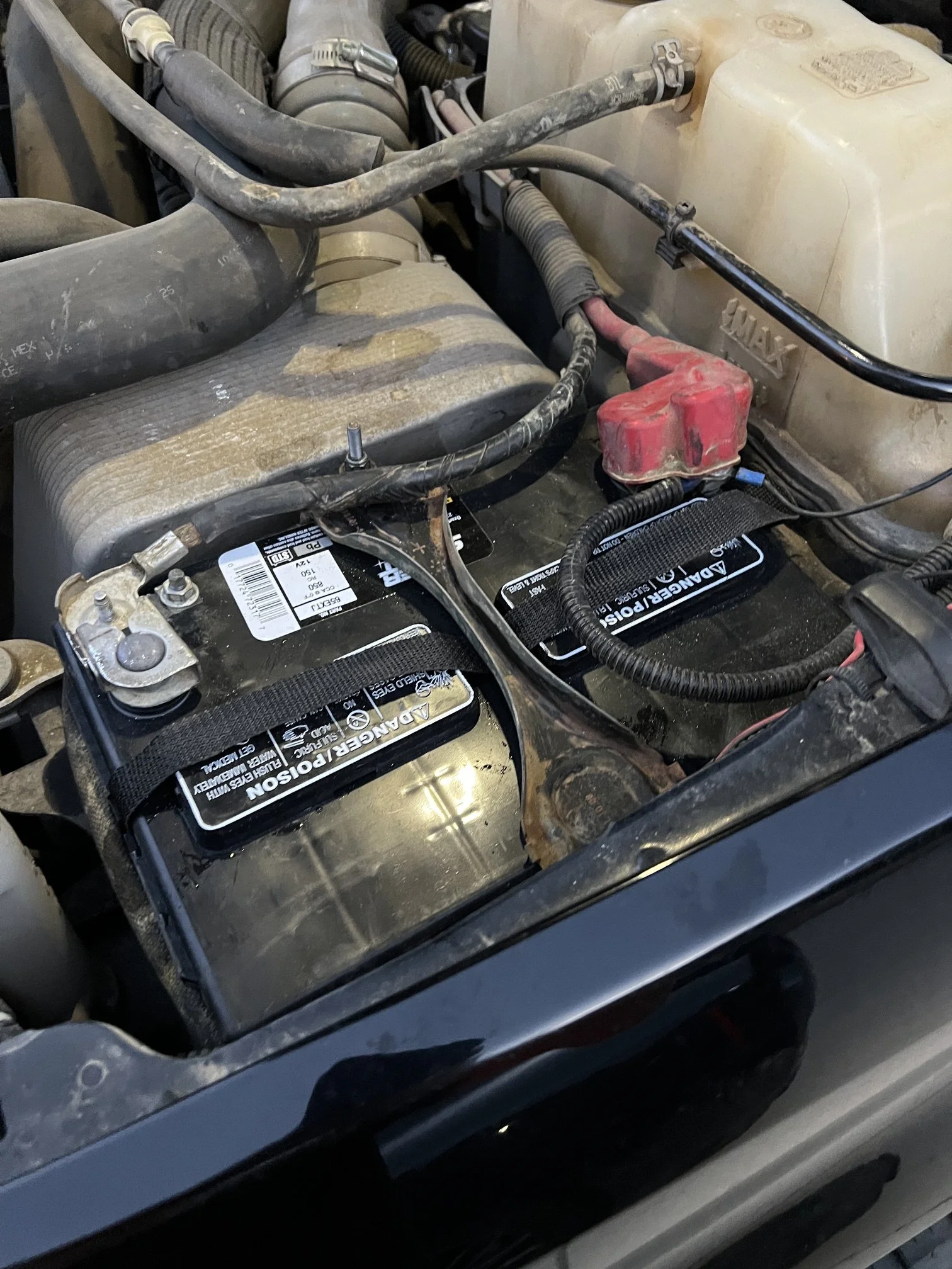 The image shows the engine compartment of a vehicle, with a focus on the battery, coolant reservoir, and various hoses and wiring. The battery appears to be a 12V lead-acid type with some dust and dirt on it.
