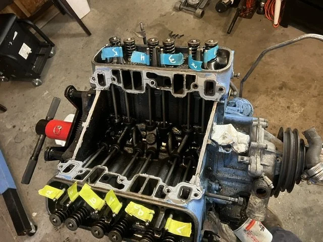 View of a disassembled engine block with valves, camshaft caps, and other components, situated on a workshop floor.