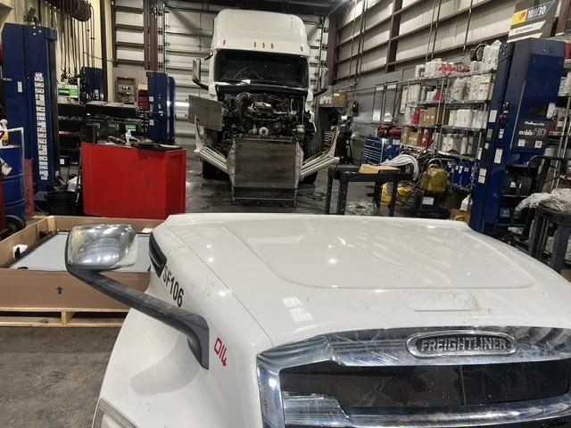 Inside an industrial garage with an unfinished truck in the background and a large white truck cab in the foreground.