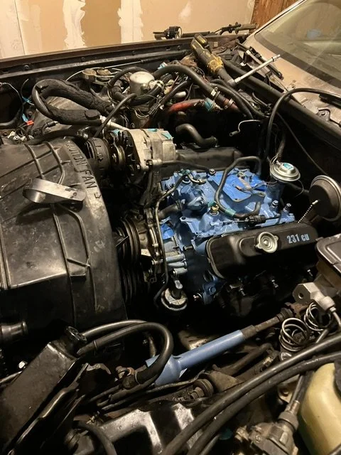 Close-up of a car engine bay showing an inline-six engine with blue engine block and various hoses and components.
