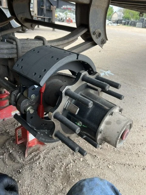A vehicle's axle with a mounted brake rotor, caliper, and a dial indicator for measuring runout, on a dirt surface with a blurred background of a trailer and trees.