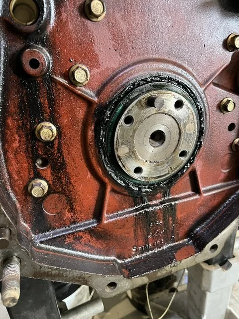 Close-up of a red engine block with a central circular metal component, surrounded by multiple gold-colored bolts and black grease or oil stains.