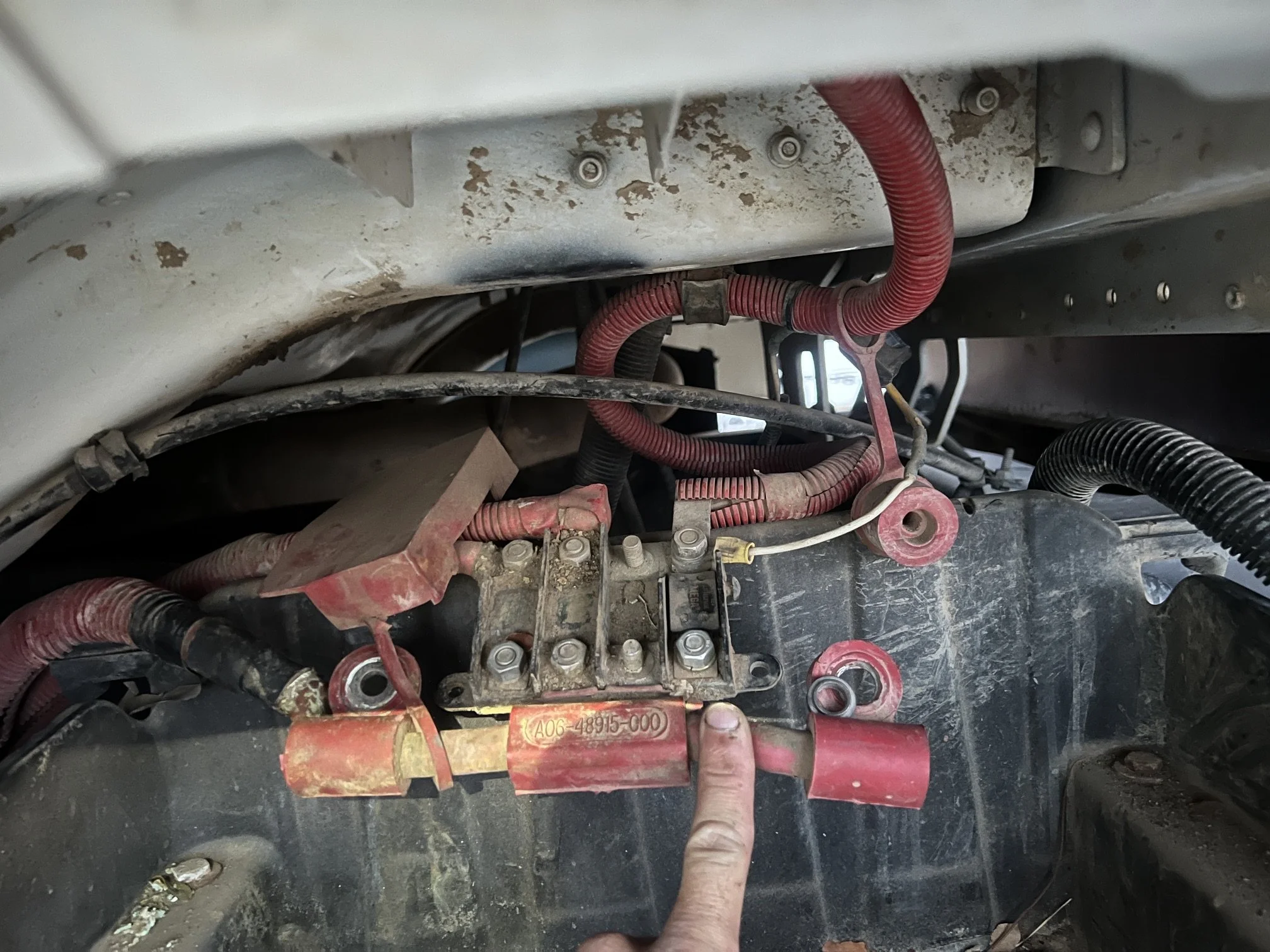 Close-up of electrical wiring and components inside a machine or vehicle with a person's finger pointing at a part, showing various red and black wires and metal parts, some with labels.