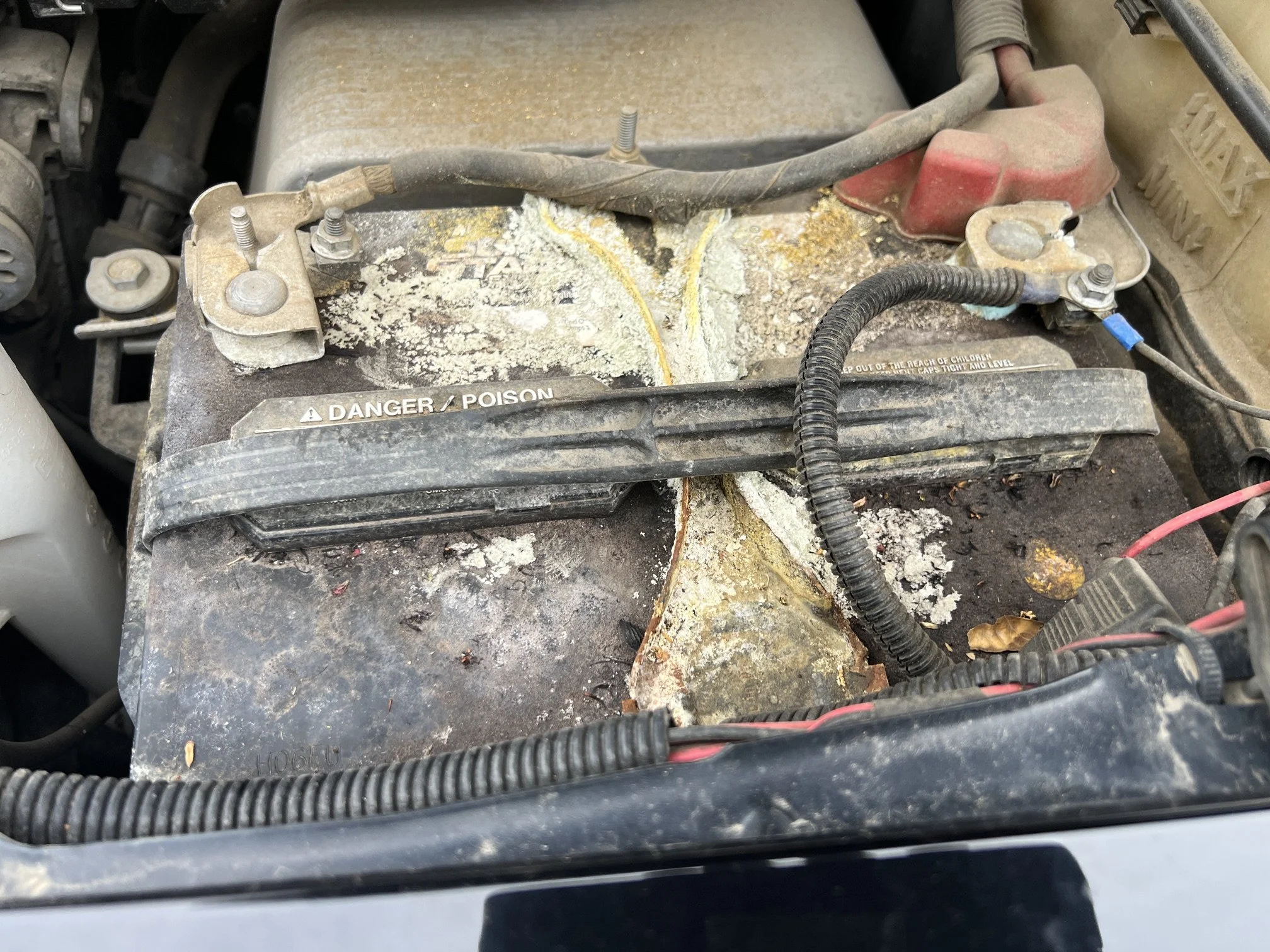 Dirt and debris on a car battery with corrosion buildup and worn labels, surrounded by various engine components and cables.