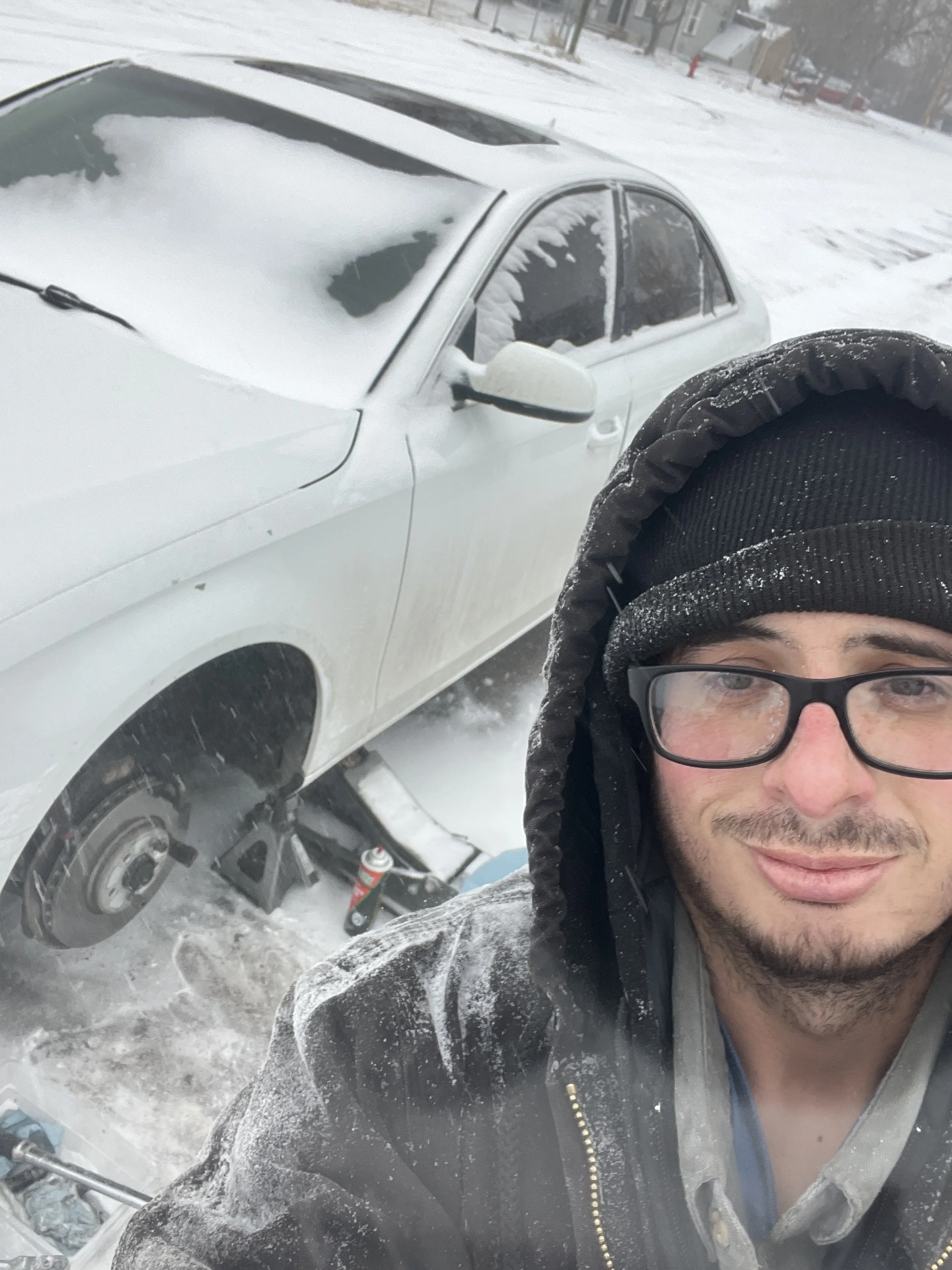 A person wearing glasses, a hooded jacket, and snow on their face taking a selfie outdoors next to a snow-covered car on a snowy day.