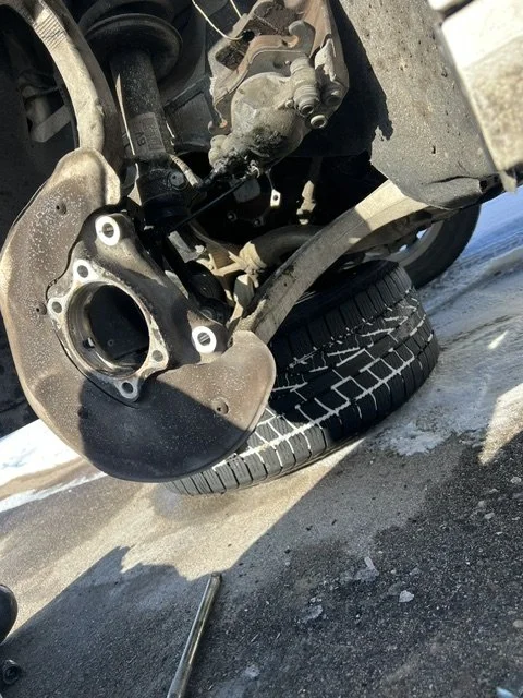 Close-up of a car's front wheel removed, showing brake caliper and suspension components, with the tire resting on the ground.