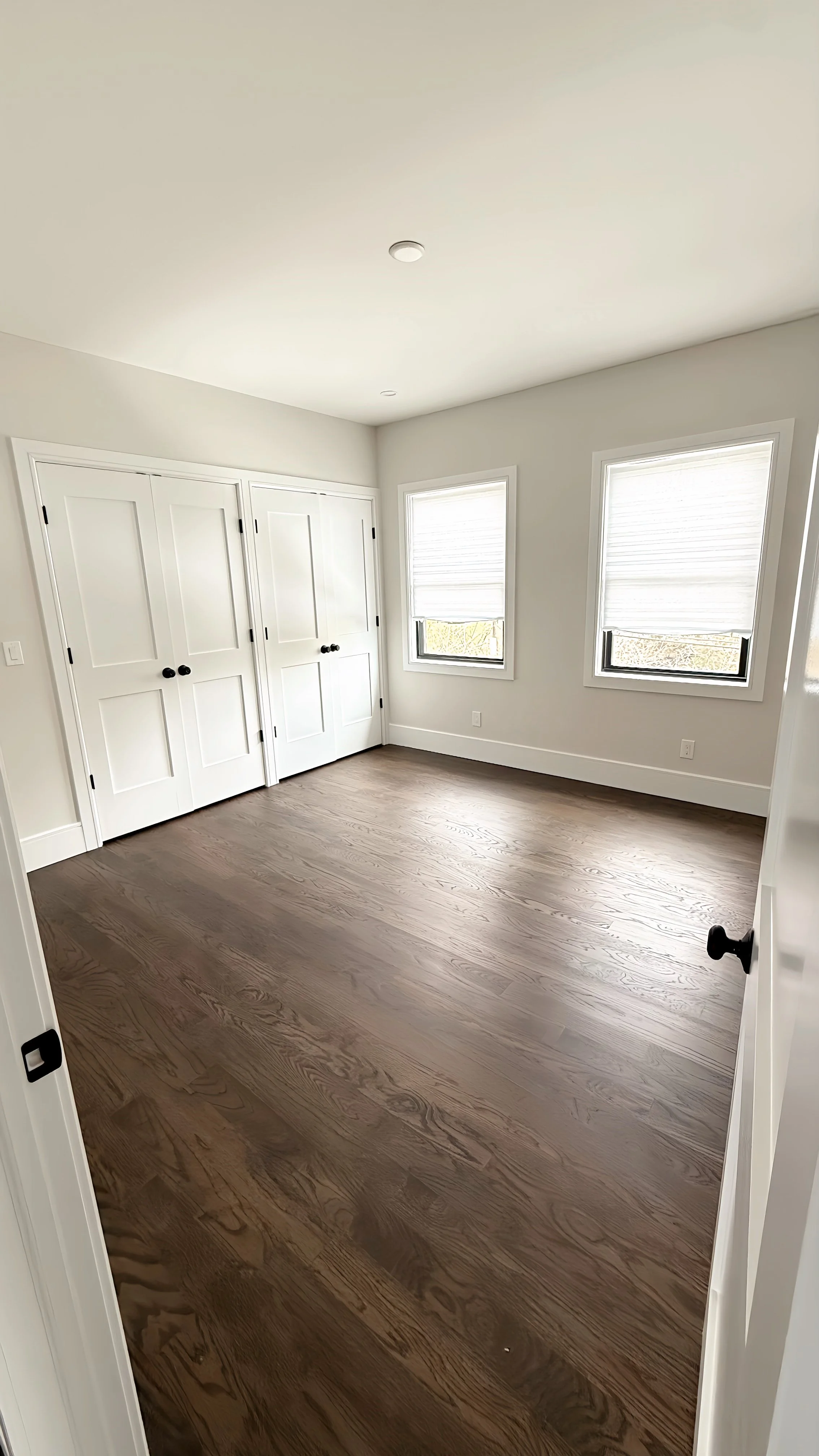 An empty room with white walls, two windows with white blinds, hardwood floors, and white double closet doors.