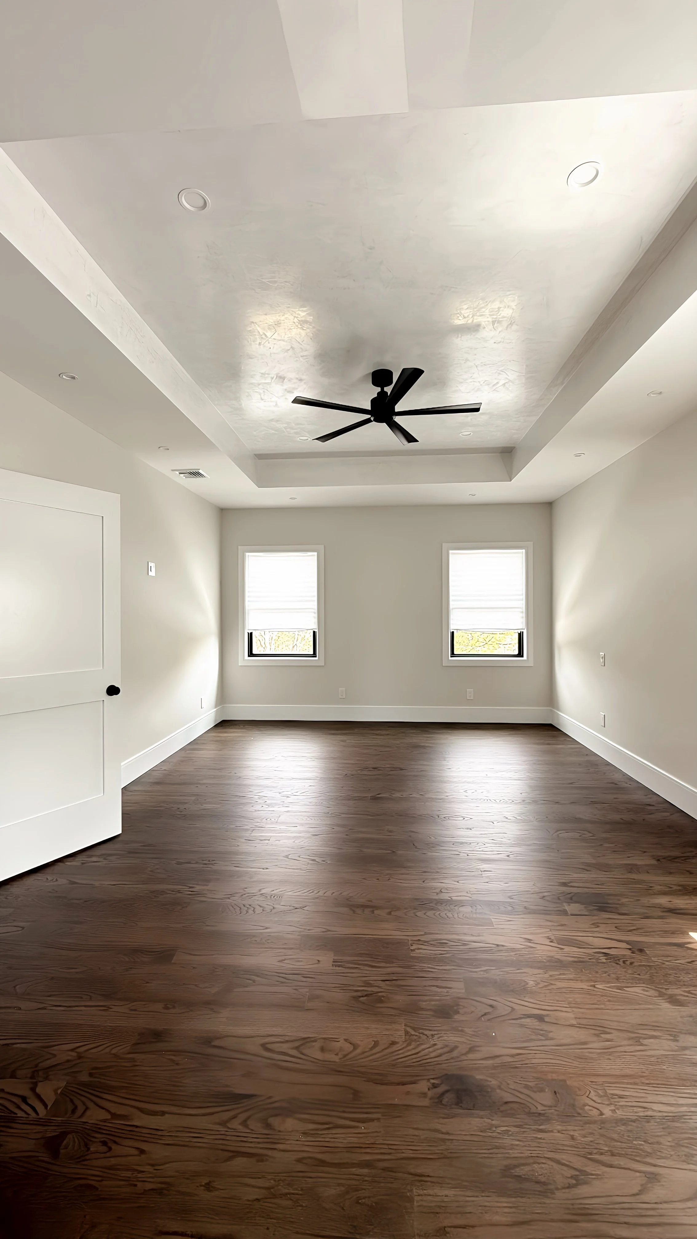 Empty room with two windows, a white ceiling with a black ceiling fan, hardwood floors, and white walls.