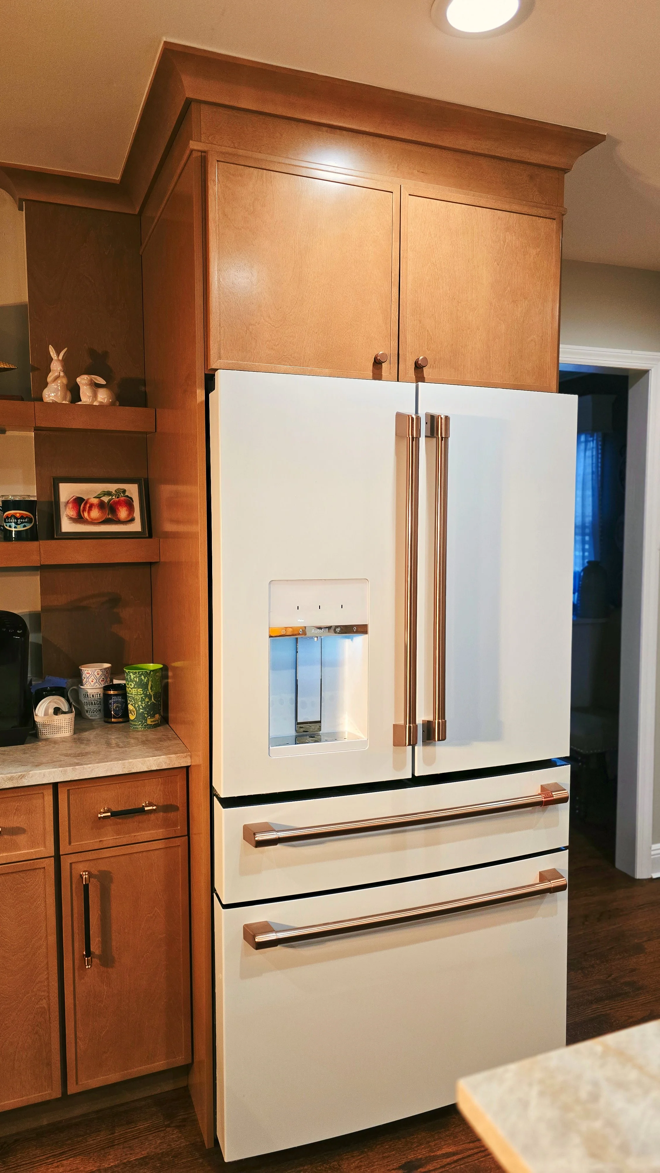 A modern kitchen with wooden cabinets and a white refrigerator that has gold-colored handles. The refrigerator features a water and ice dispenser on the door. To the left, there is a countertop with mugs and small decorative items, including rabbit f