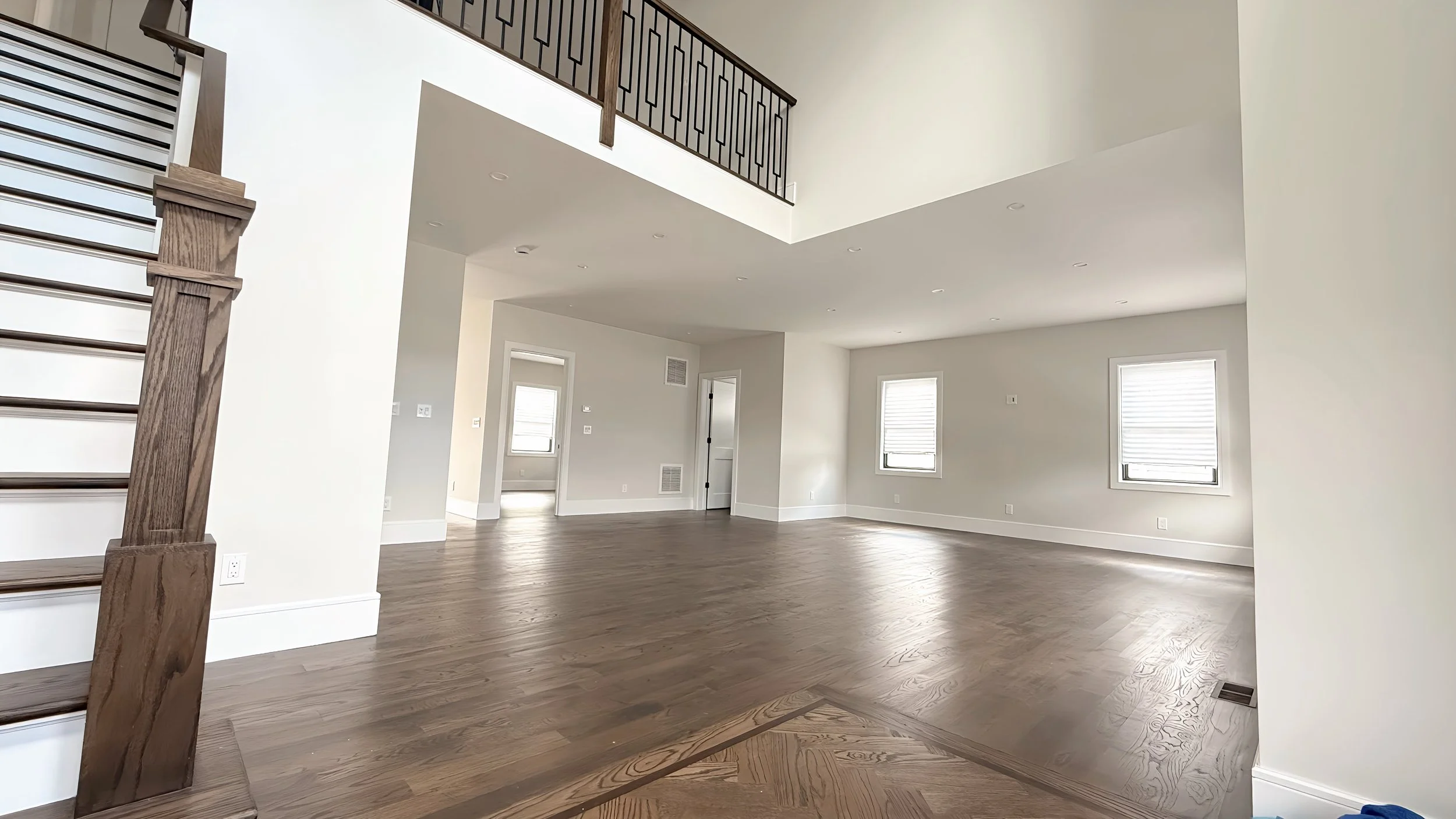 Empty living room with white walls, hardwood floors, three windows, and a staircase with a wooden banister on the left.