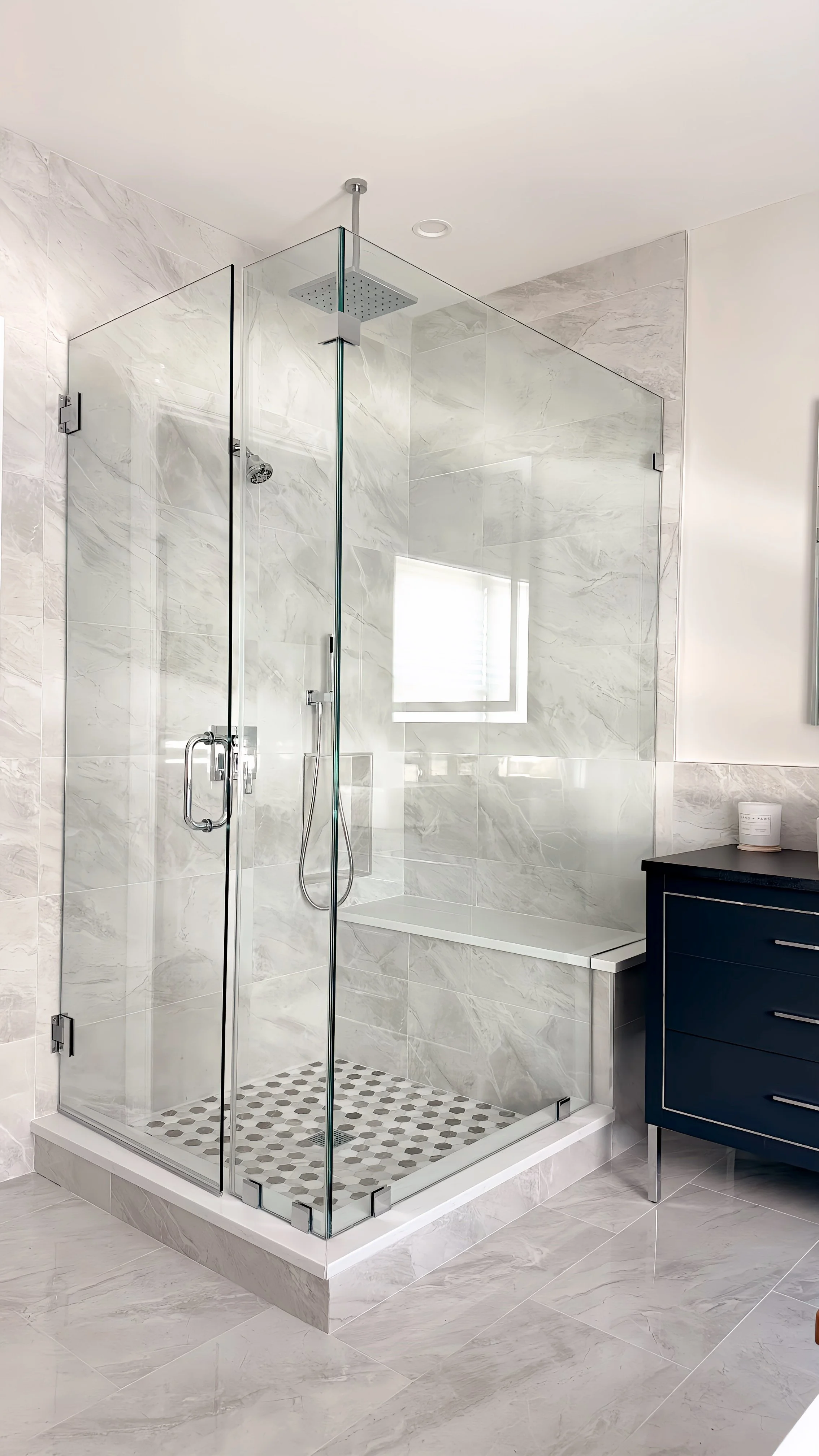 Modern bathroom with glass shower enclosure, white marble tile walls, and a black vanity with drawers. The shower has a rainfall showerhead and a handheld shower, with gray and white patterned floor tiles.