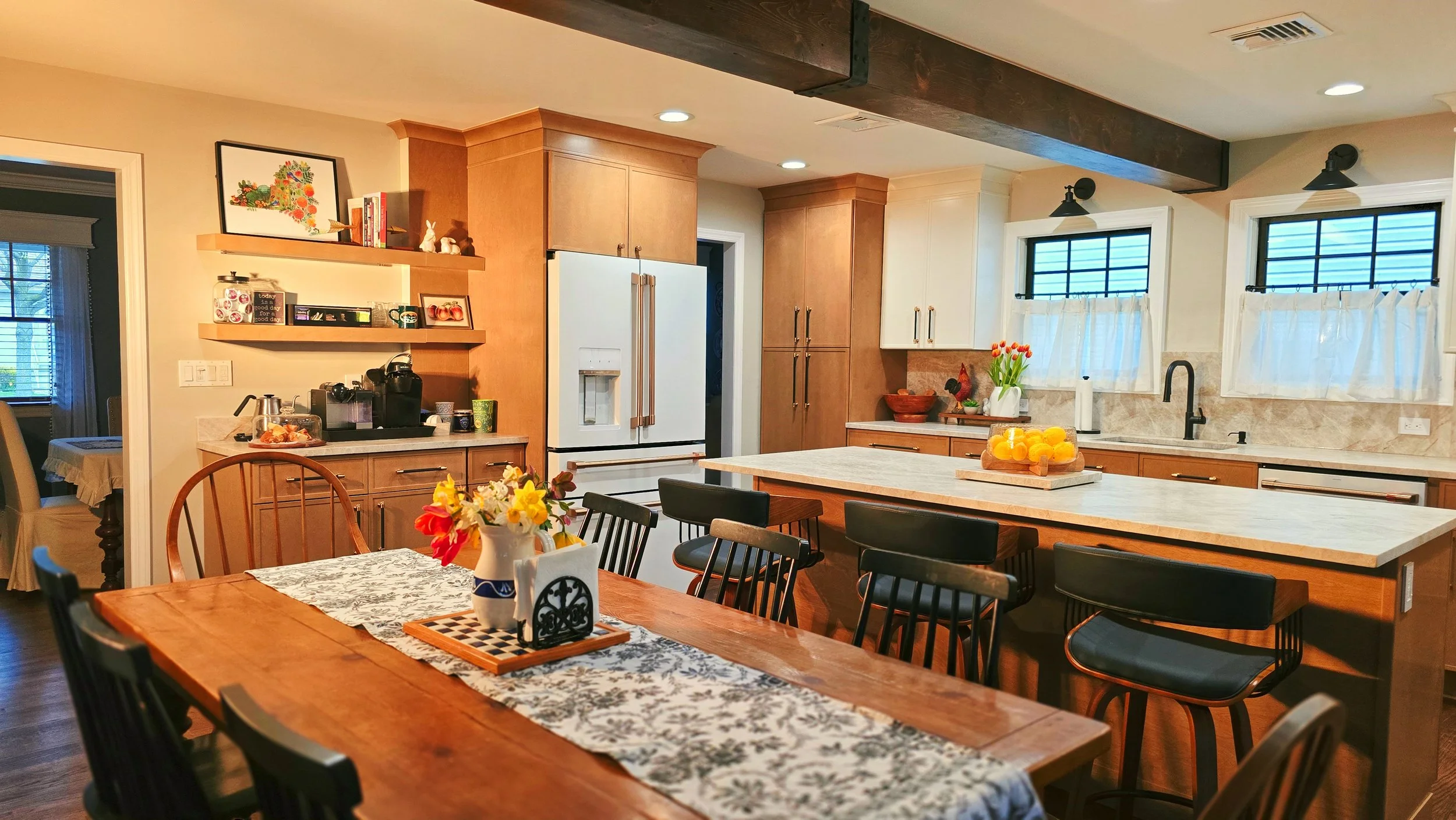 A cozy kitchen with a large dining table decorated with a floral table runner and a centerpiece of colorful flowers. The kitchen features wooden cabinets, a white refrigerator, a marble countertop, and two windows with white curtains. There are various kitchen items and decorations, including a fruit bowl with oranges and a small vase with tulips.
