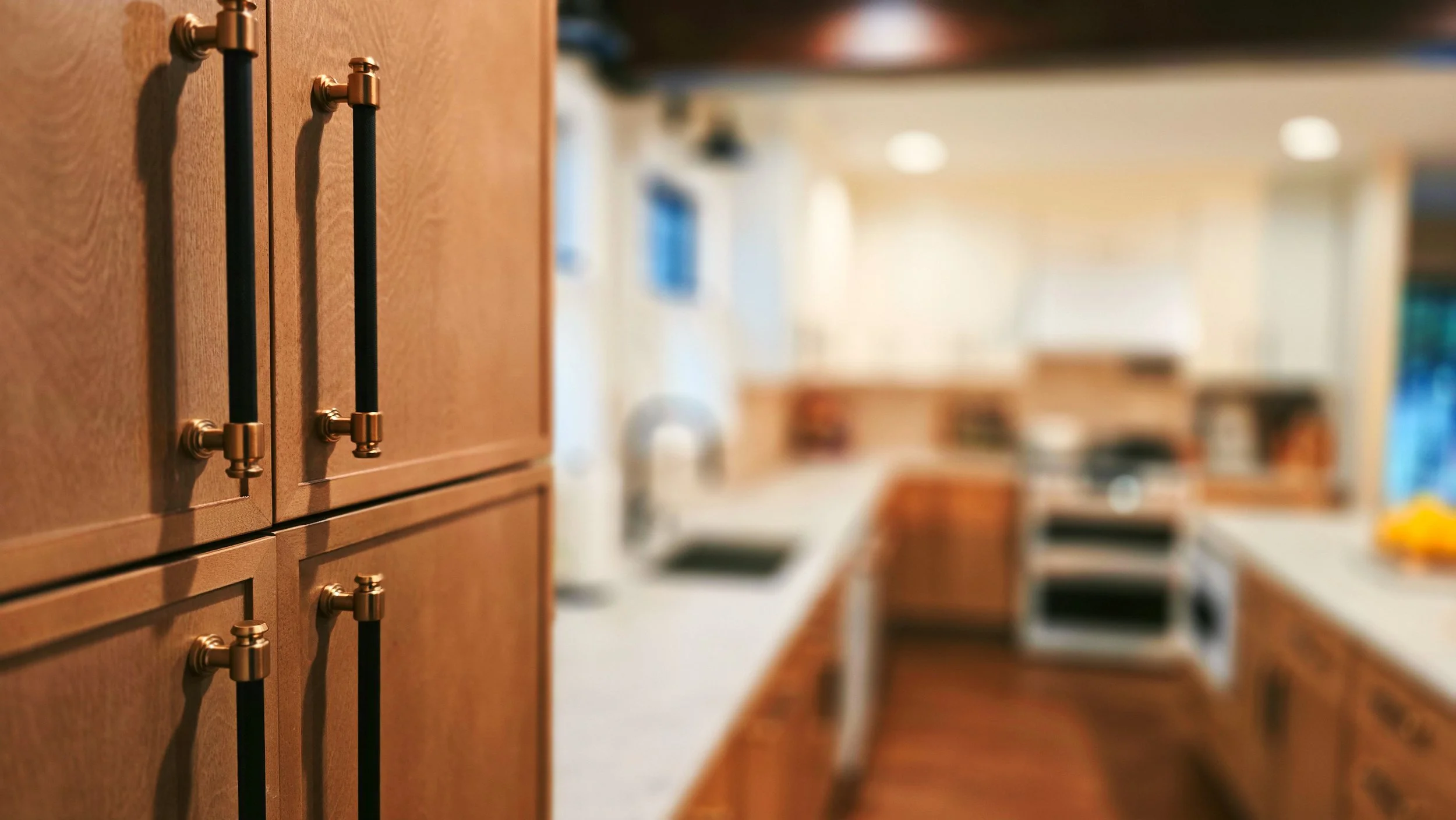 Close-up of a wooden kitchen cabinet with black and gold metal handles, blurring into a bright kitchen background.