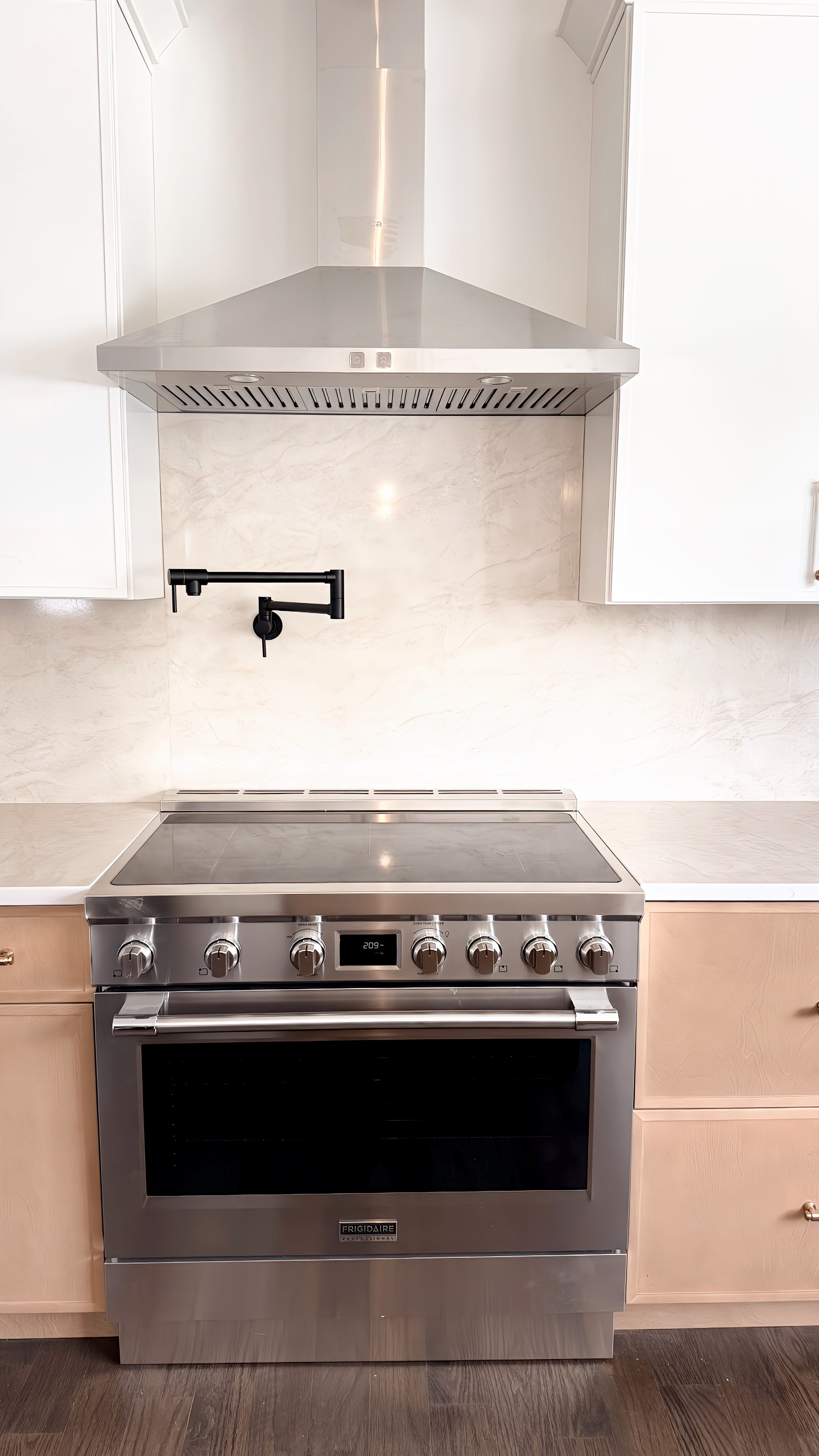 Stainless steel oven and stovetop with black control knobs, beige cabinets on either side, white backsplash, and a black faucet above the stove in a modern kitchen.