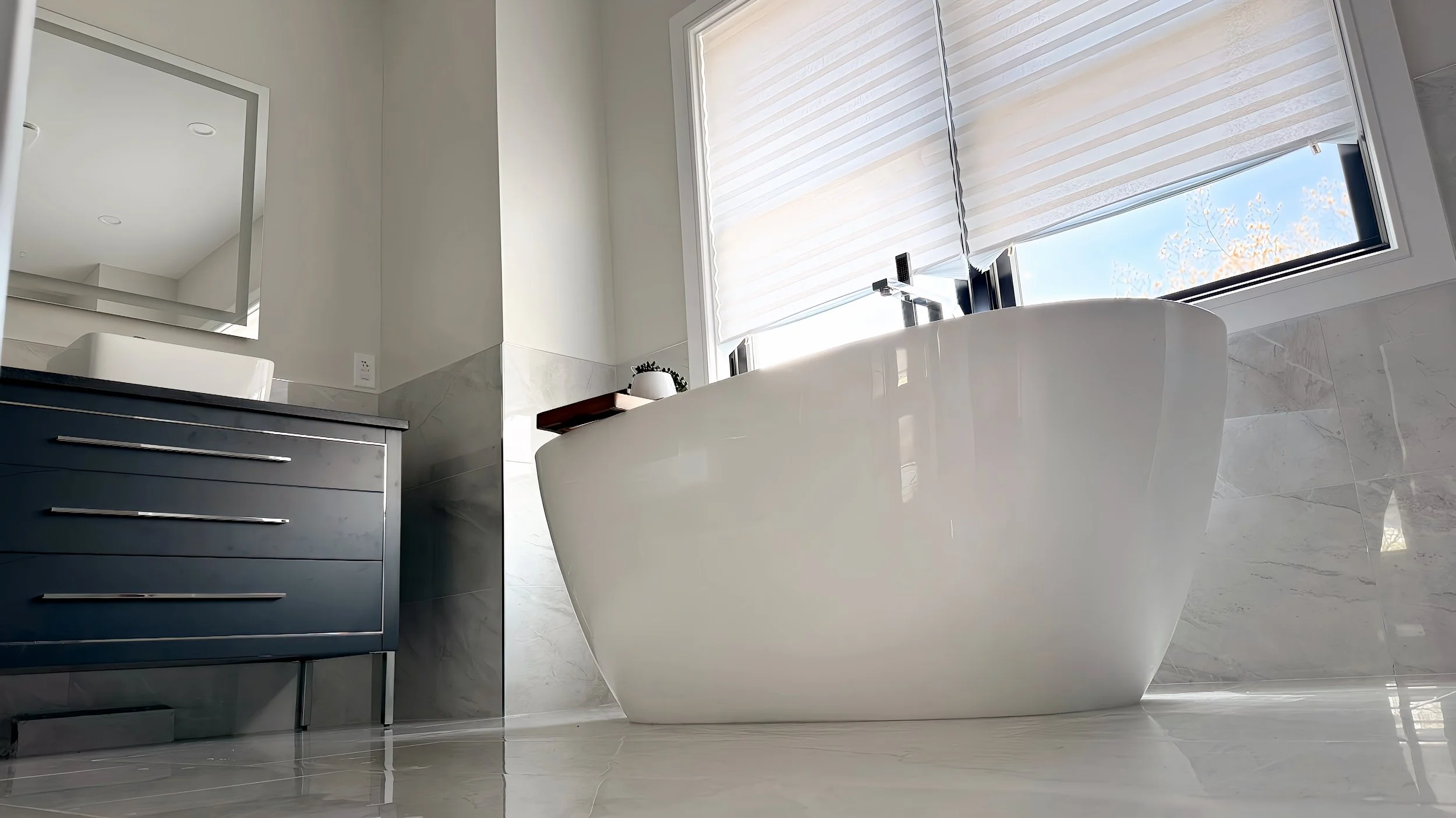 Modern bathroom with a white freestanding bathtub, large window with horizontal blinds, gray dresser, and a mirror on the wall.