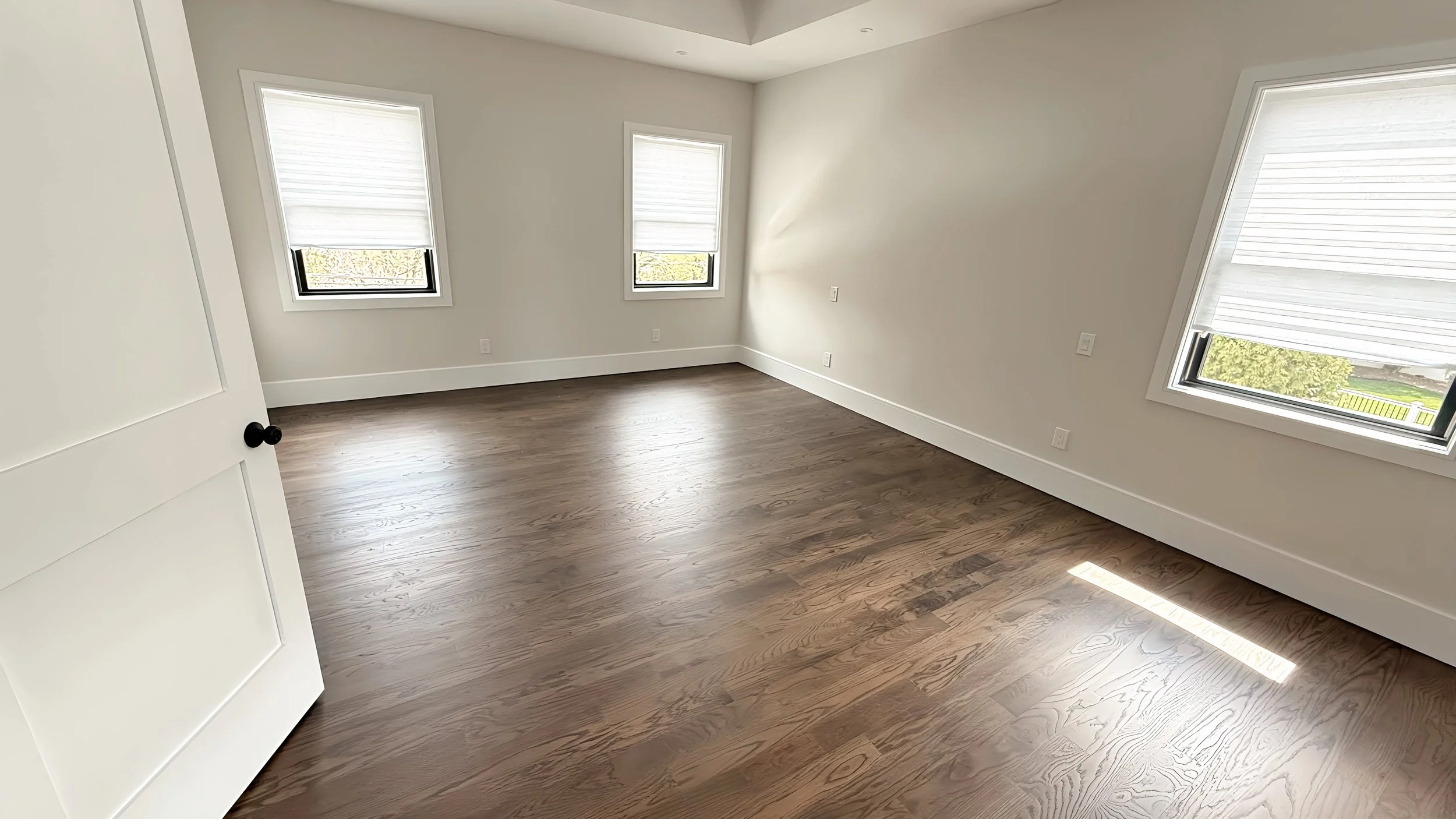 Empty room with white walls, hardwood floors, and three windows with white blinds letting in natural light.