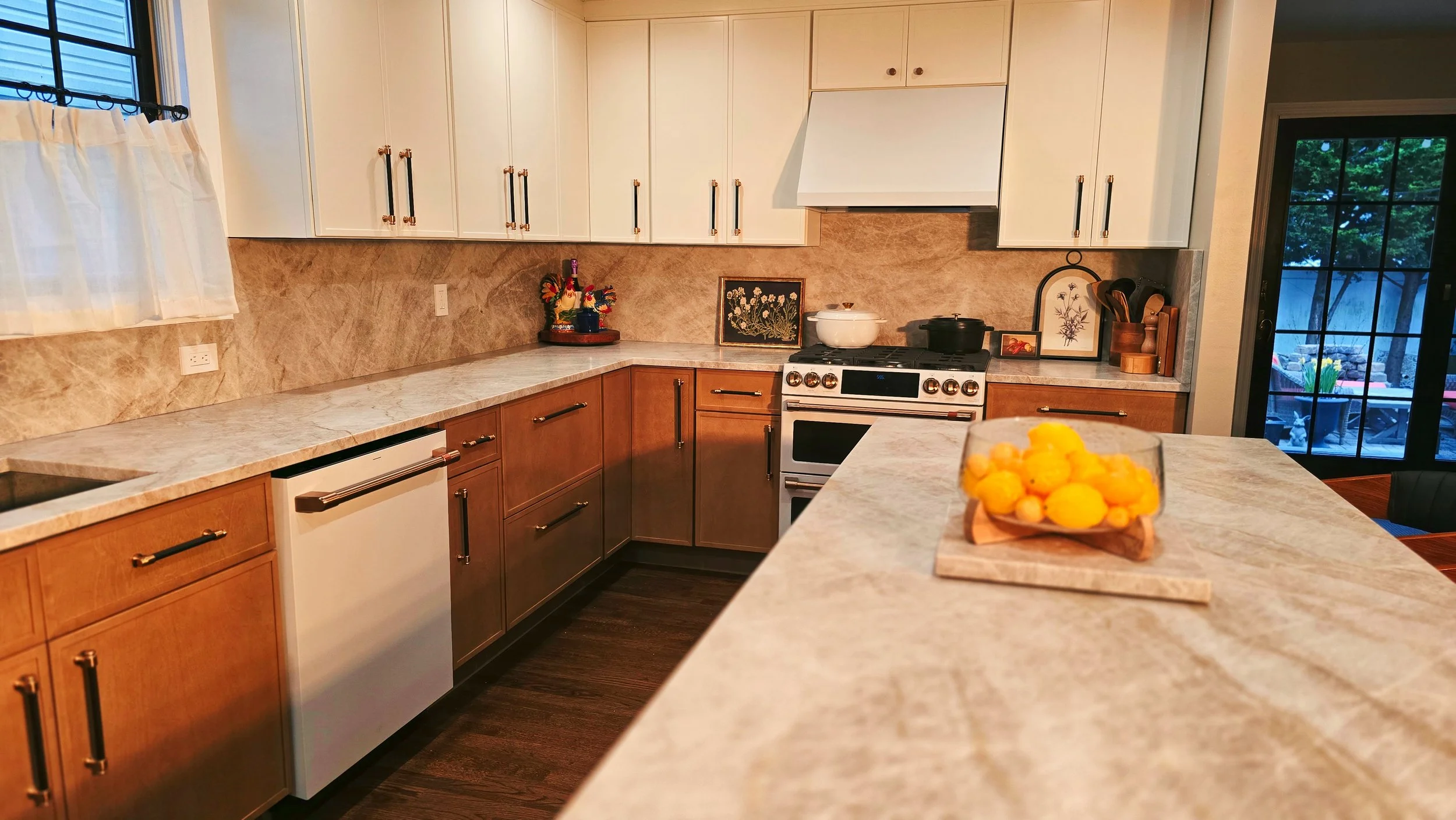 A kitchen with beige and white cabinets, a marble countertop, and a sliding glass door. There is a bowl of lemons on the island in the foreground, and several decorative items and appliances on the countertops.