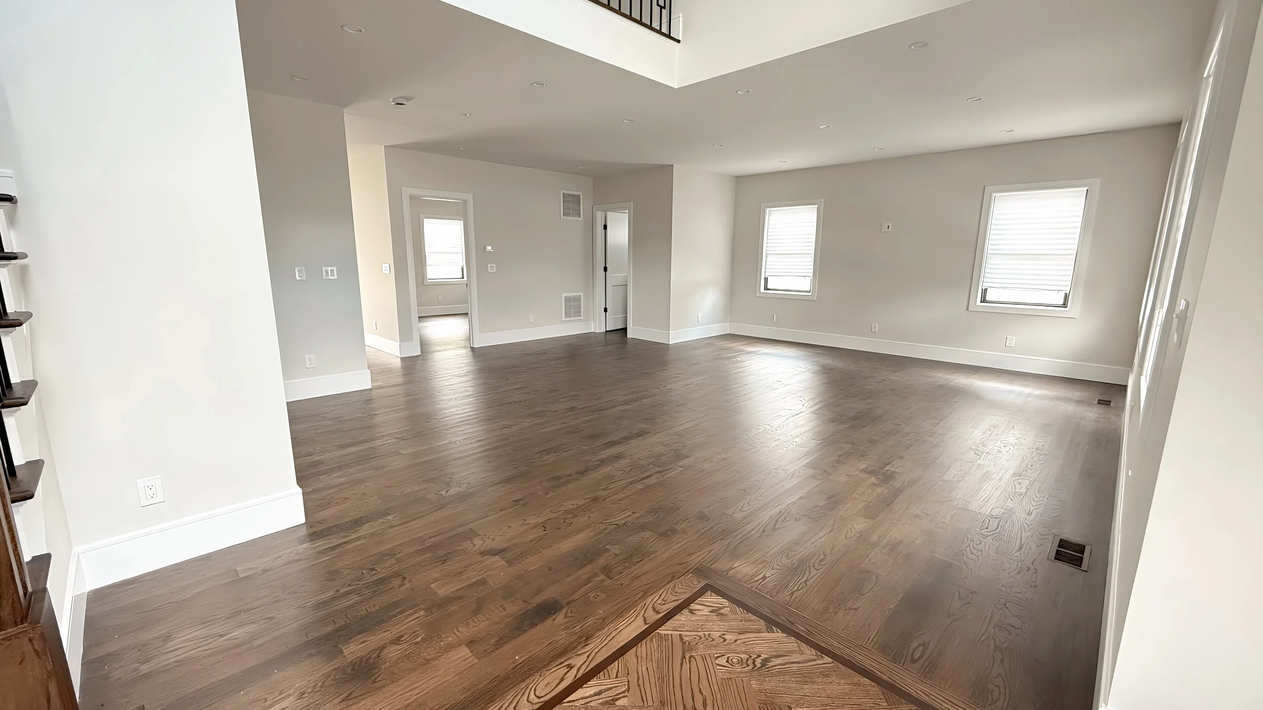 Empty living room with hardwood floors, white walls, multiple windows with blinds, and recessed lighting.