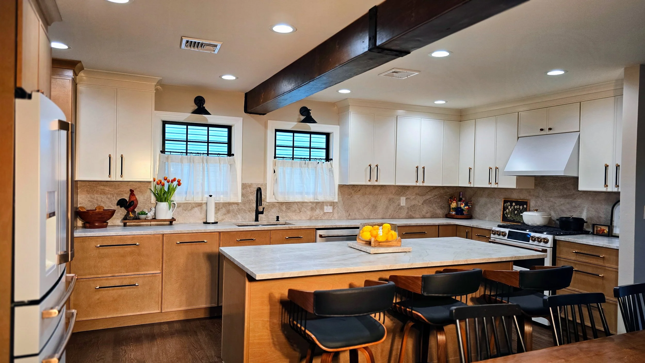 A modern kitchen with white cabinets, two small windows with white curtains, a marble countertop island with lemons, and a table with black chairs.